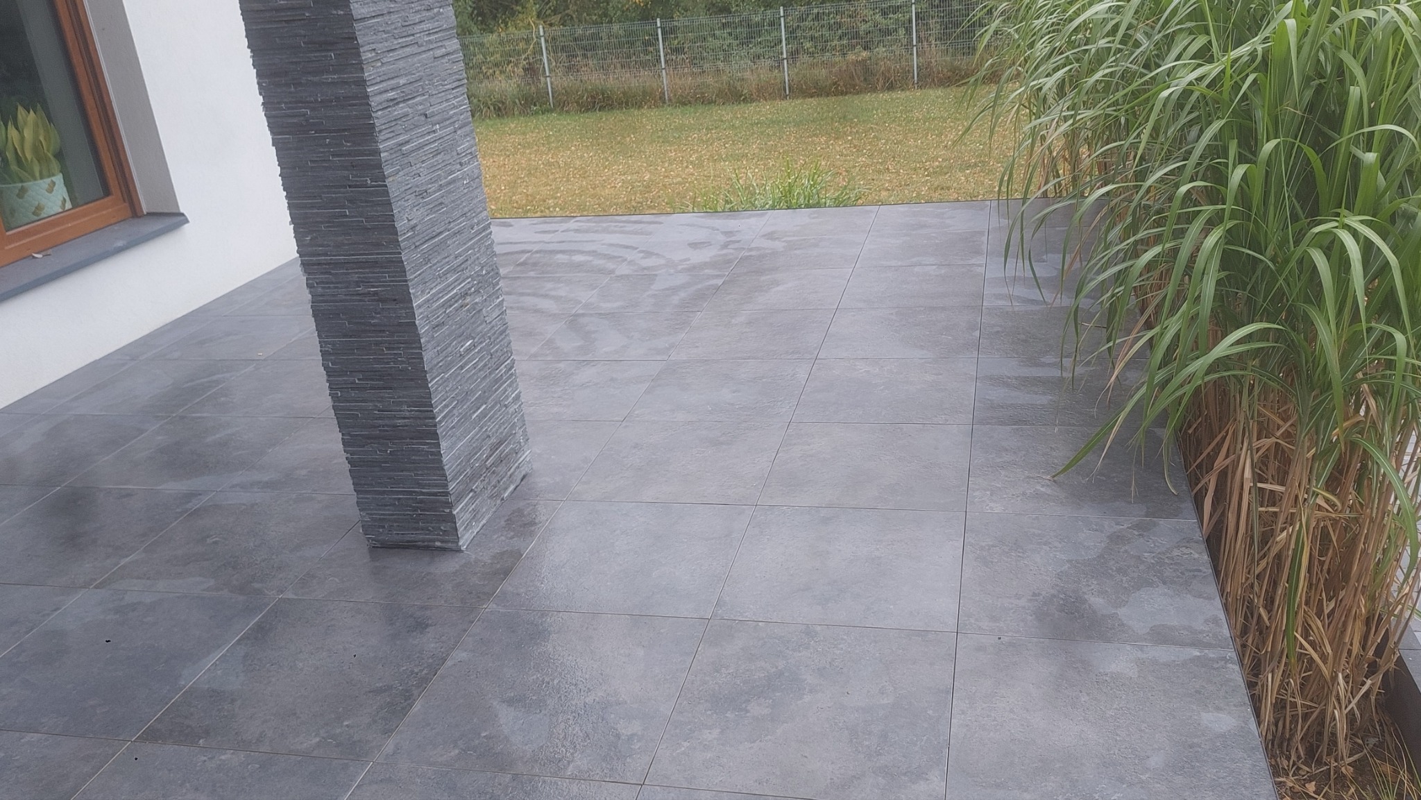 Szare płytki na tarasie z dekoracyjnym słupem z kamienia łupkowego i ozdobną trawą, widok na trawnik za ogrodzeniem.