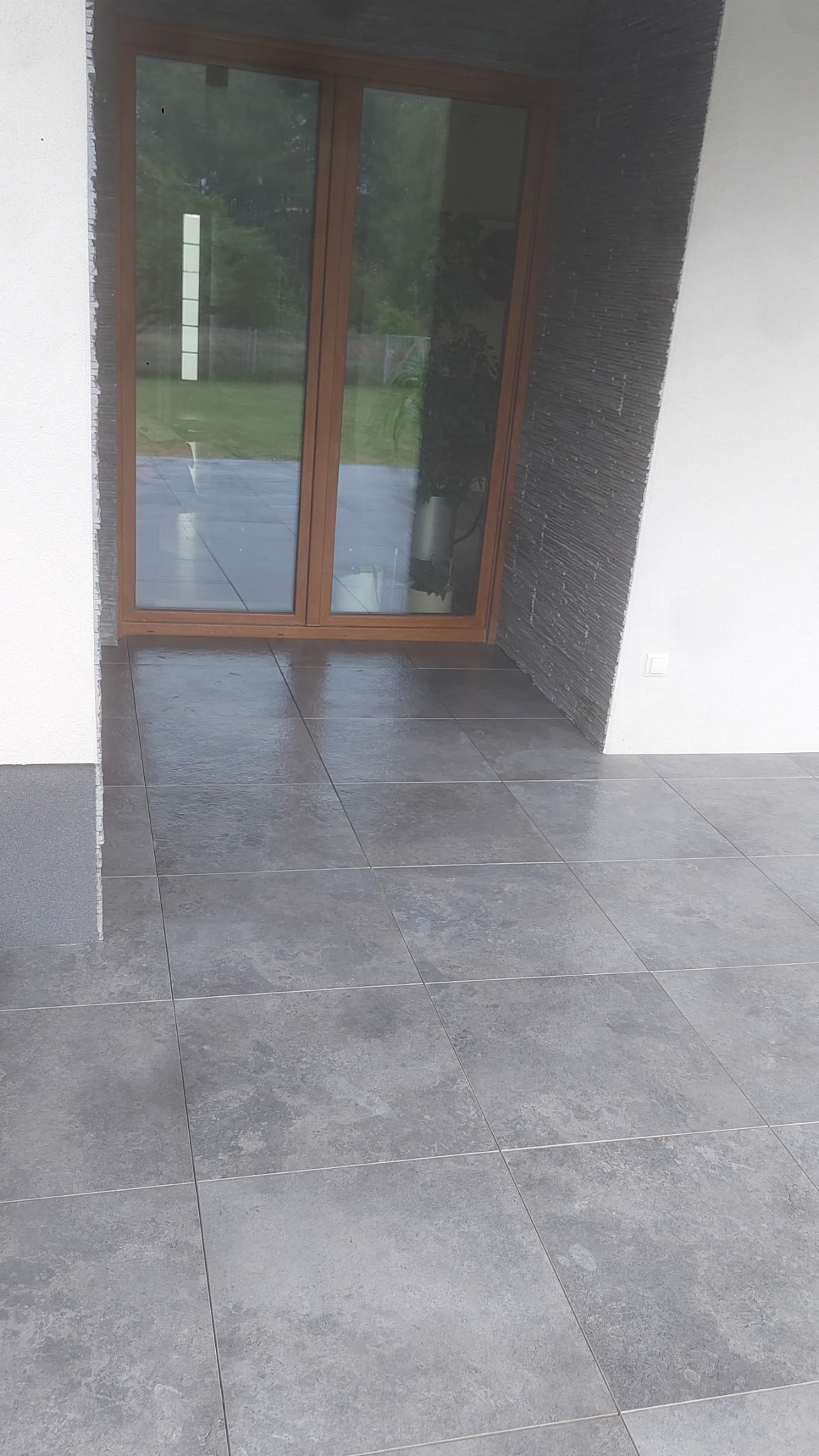 Szare płytki gresowe ułożone na zewnątrz, widoczne brązowe drzwi tarasowe z szybami i fragment elewacji z dekoracyjnym kamieniem.