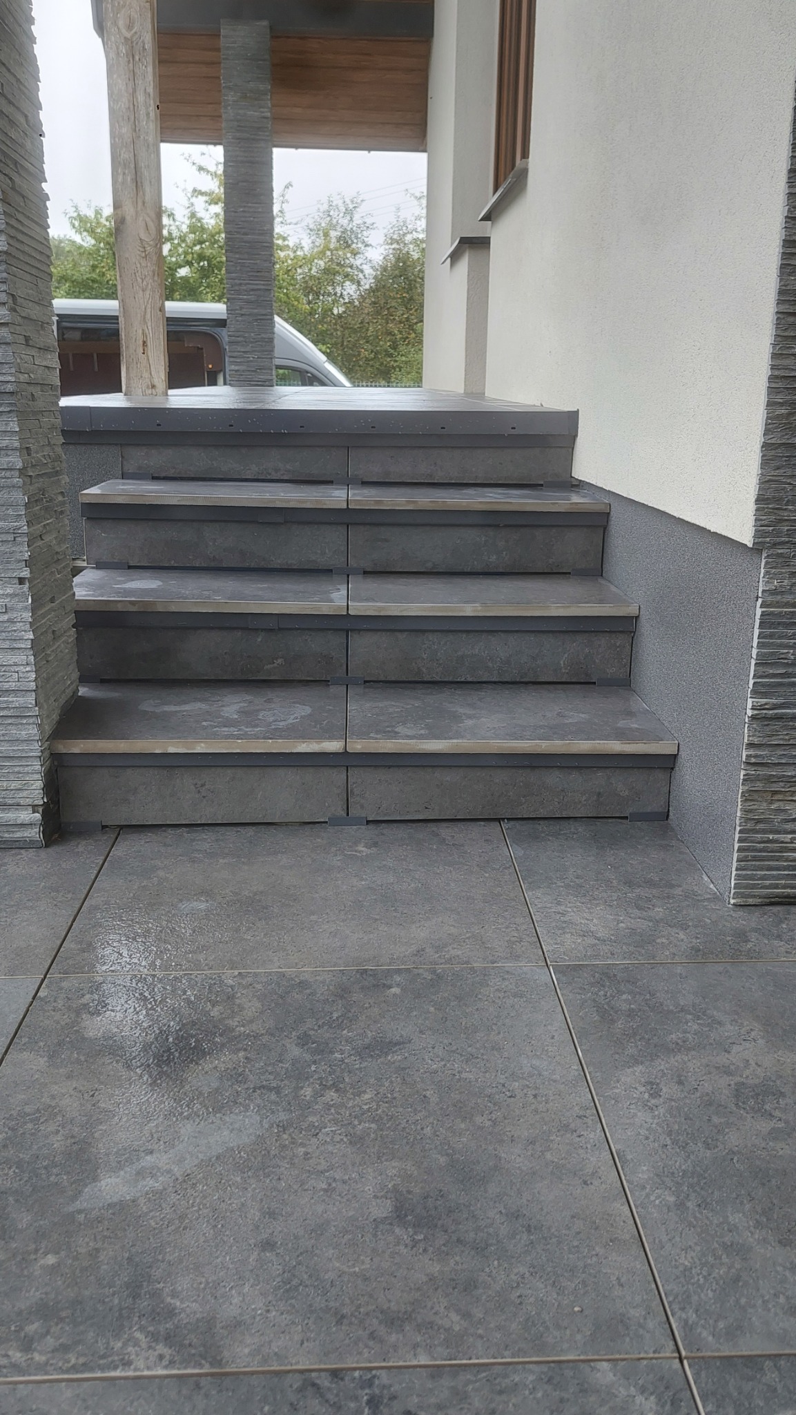Zewnętrzne schody wyłożone szarymi płytkami, z widocznymi fugami i fragmentem elewacji budynku. Słupki wykończone dekoracyjnym kamieniem. Lekko pochmurne niebo w tle.