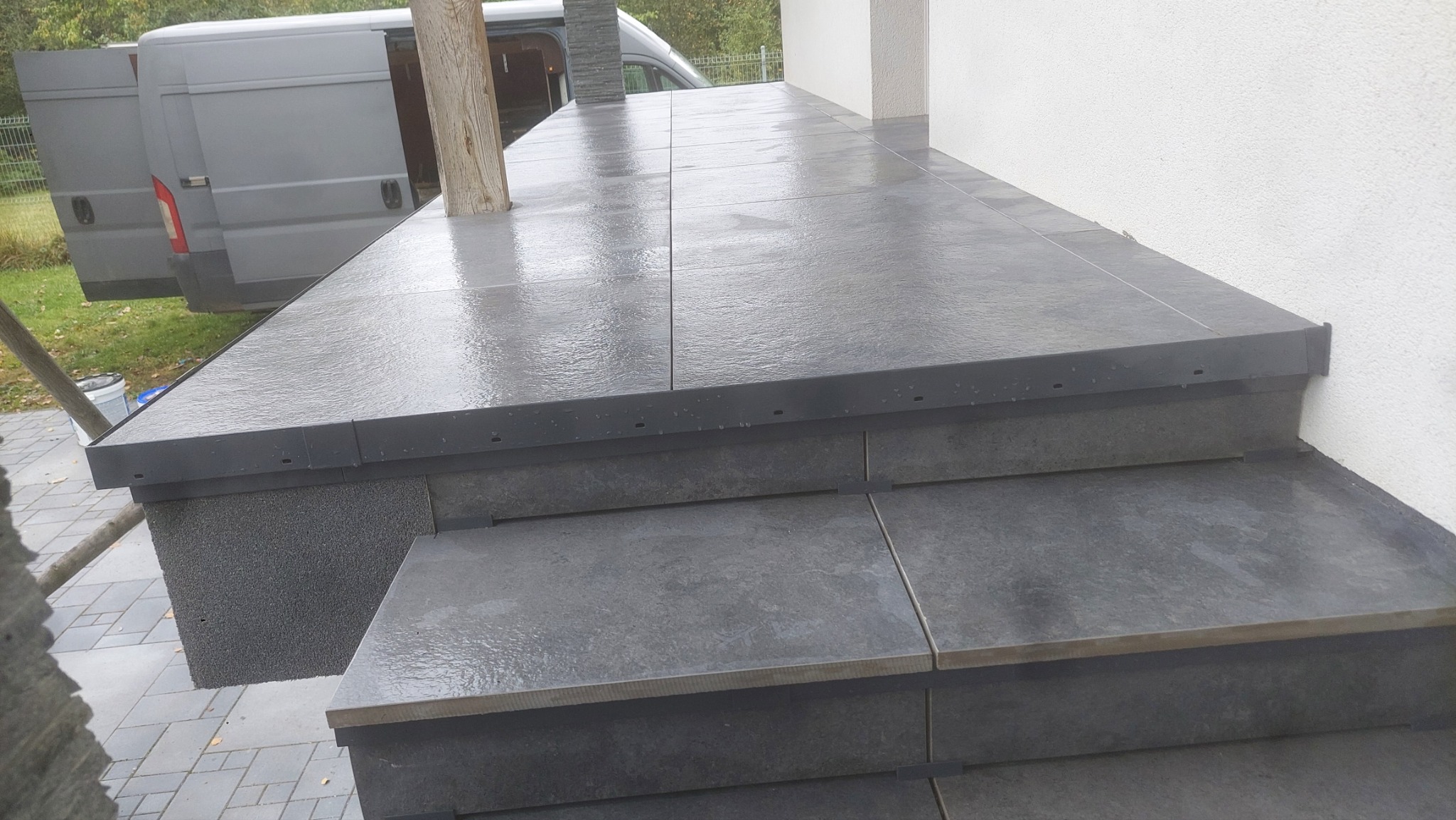 Zewnętrzne schody i taras wyłożone szarą płytką ceramiczną, widoczny fragment zaparkowanego busa w tle.