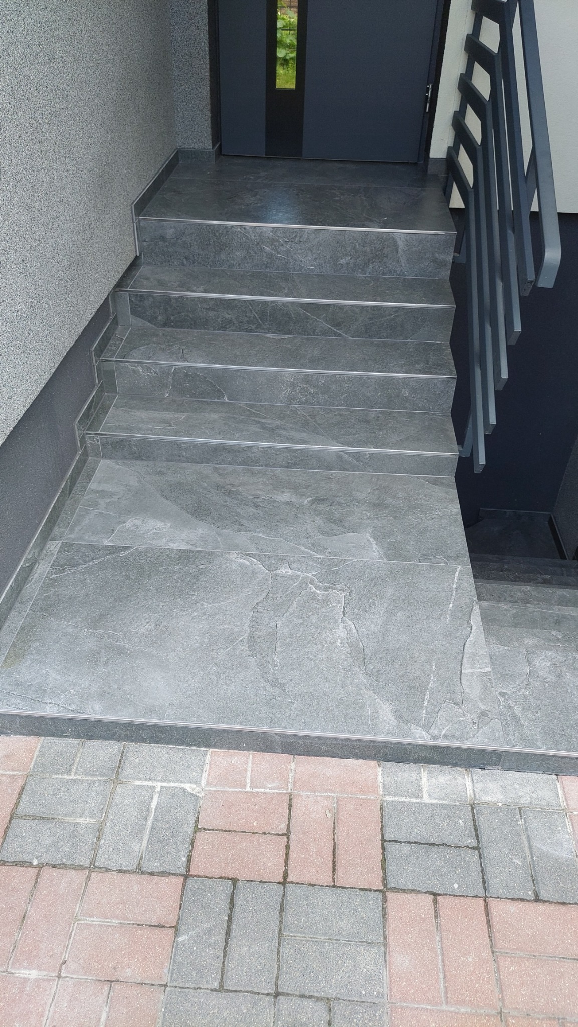 Zewnętrzne schody wyłożone szarą płytką imitującą kamień, z metalową poręczą i wejściem do budynku z szarą stolarką.