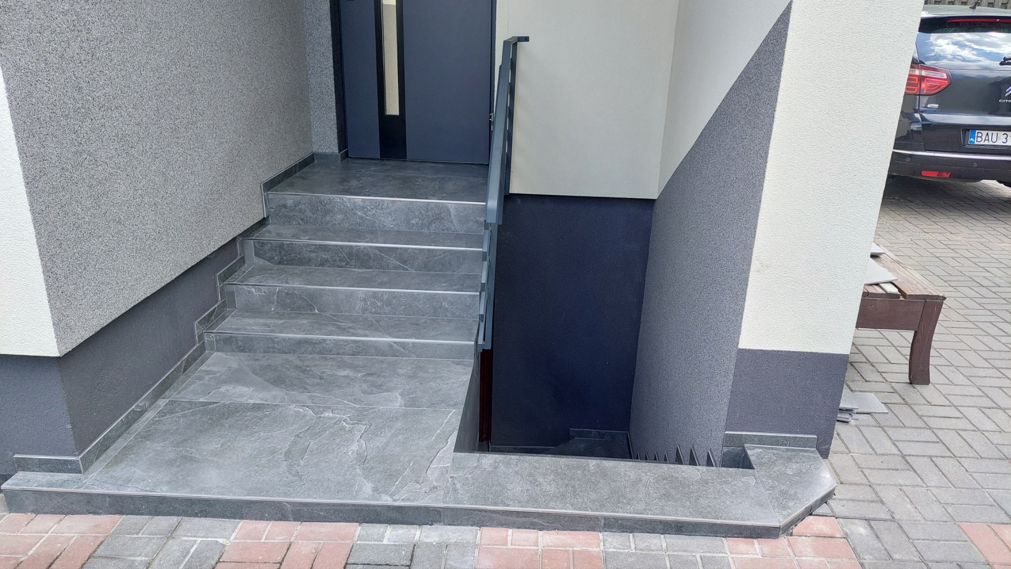 Zewnętrzne schody wejściowe wyłożone szarymi płytkami imitującymi marmur, z metalową barierką, prowadzące do ciemnego wejścia. Widoczny fragment elewacji budynku w dwóch odcieniach szarości i bieli...