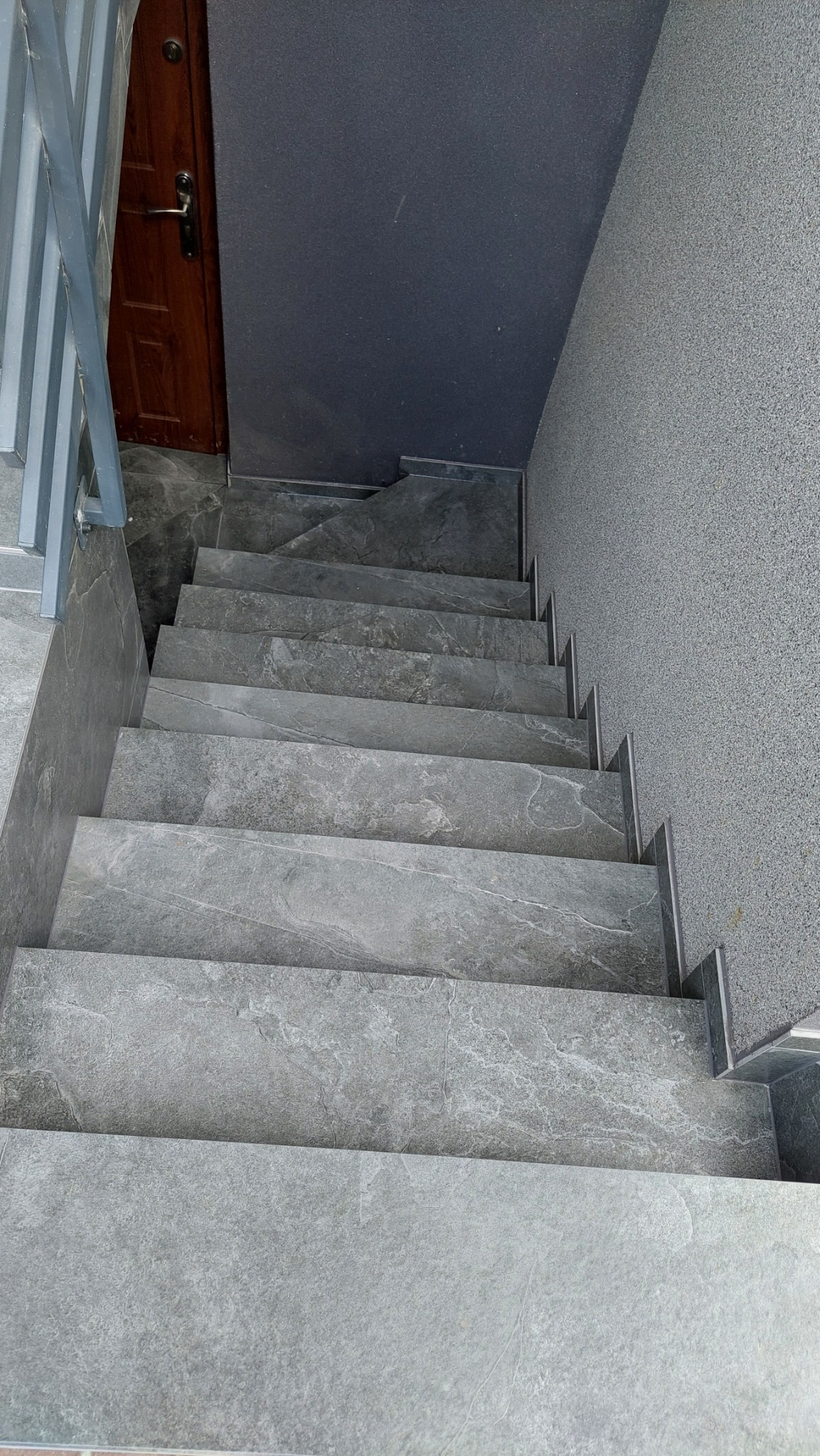 Schody wyłożone szarymi płytkami imitującymi kamień, widok z góry, z drewnianymi drzwiami na górze i szarą, teksturowaną ścianą po prawej stronie.