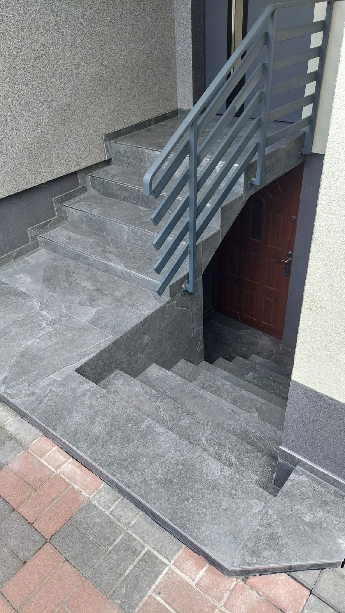 Zewnętrzne schody wyłożone szarą płytką imitującą kamień, z metalową balustradą, prowadzące do brązowych drzwi wejściowych.