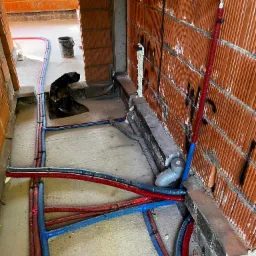 Wnętrze budynku w trakcie budowy, widoczne prowadzone instalacje wodne i kanalizacyjne w kolorze niebieskim i czerwonym, mocowane do posadzki i ścian z cegły dziurawki. Na ścianie widoczne ślady po...