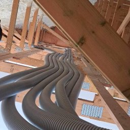 HYDROinstal Usługi Hydrauliczne Jakub Grudziński - Rzędy szarych, karbowanych rur instalacyjnych ułożone równolegle w przestrzeni poddasza z drewnianą konstrukcją i izolacją piankową.