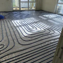 Wnętrze w trakcie prac instalacyjnych: rozłożone rury ogrzewania podłogowego na folii izolacyjnej z widocznymi oknami i fragmentem ściany z pustaków.