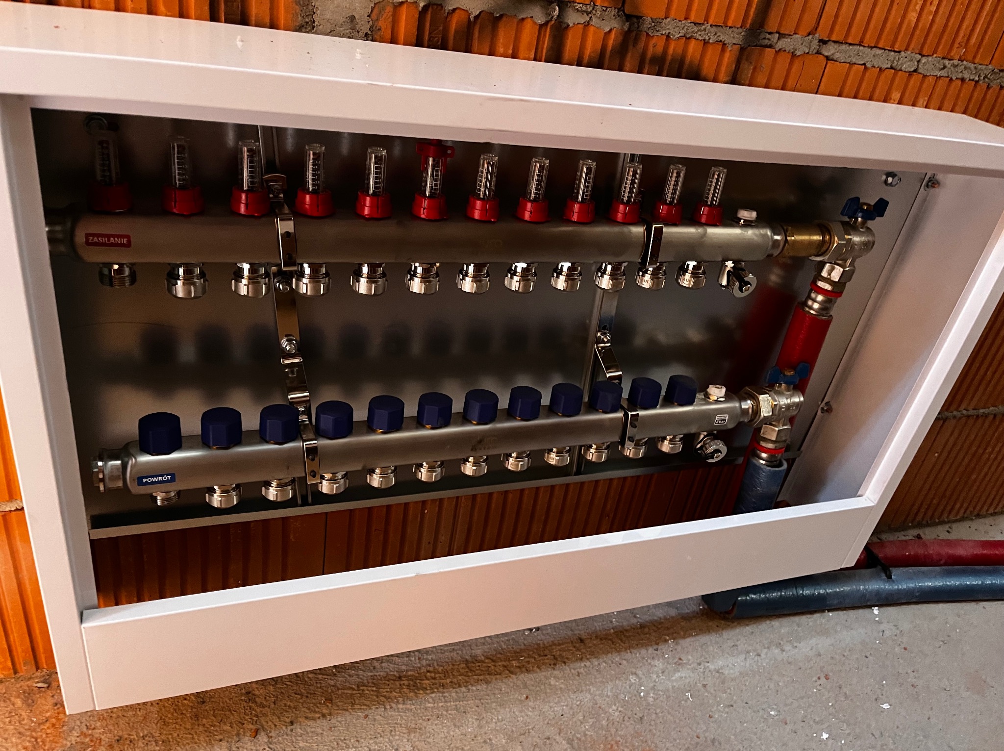 Rozdzielacz centralnego ogrzewania w białej szafce, z widocznymi rurami doprowadzającymi i odprowadzającymi wodę, zamontowany na tle ceglanej ściany.