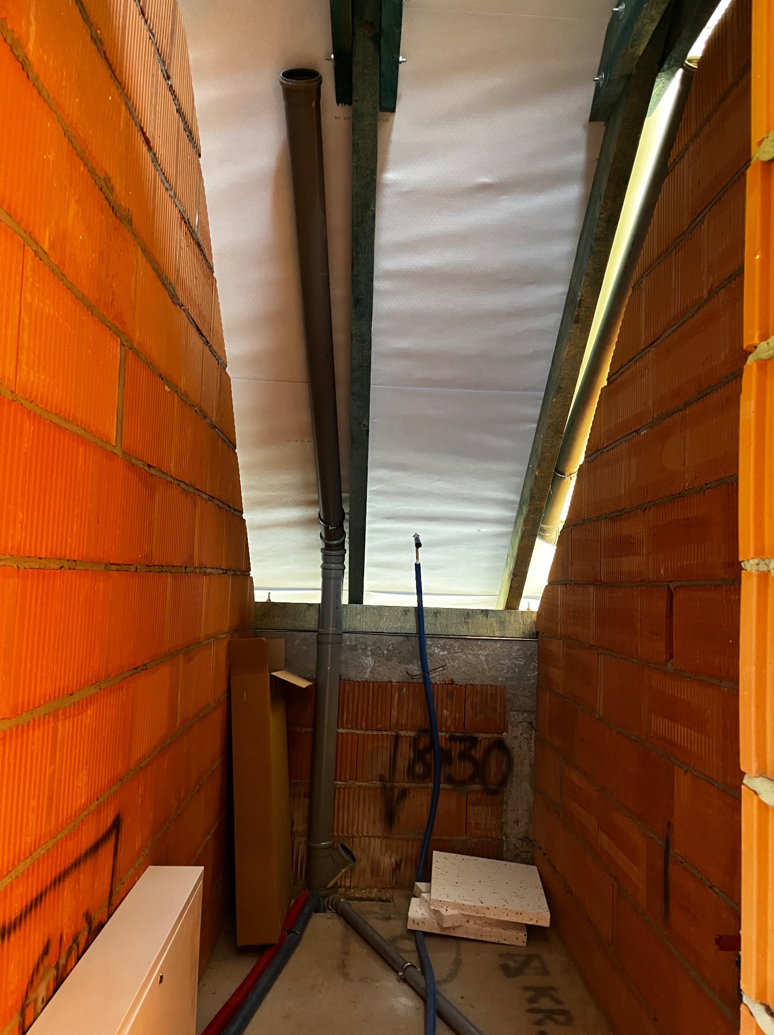 Wnętrze w trakcie budowy: widoczne ceglane ściany, rury kanalizacyjne, instalacja pod sufitem i fragmenty materiałów budowlanych na podłodze.