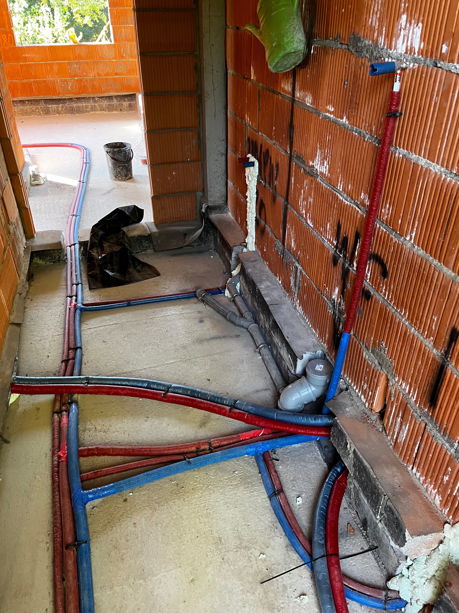 Wnętrze budynku w trakcie budowy, widoczne prowadzone instalacje wodne i kanalizacyjne w kolorze niebieskim i czerwonym, mocowane do posadzki i ścian z cegły dziurawki. Na ścianie widoczne ślady po...