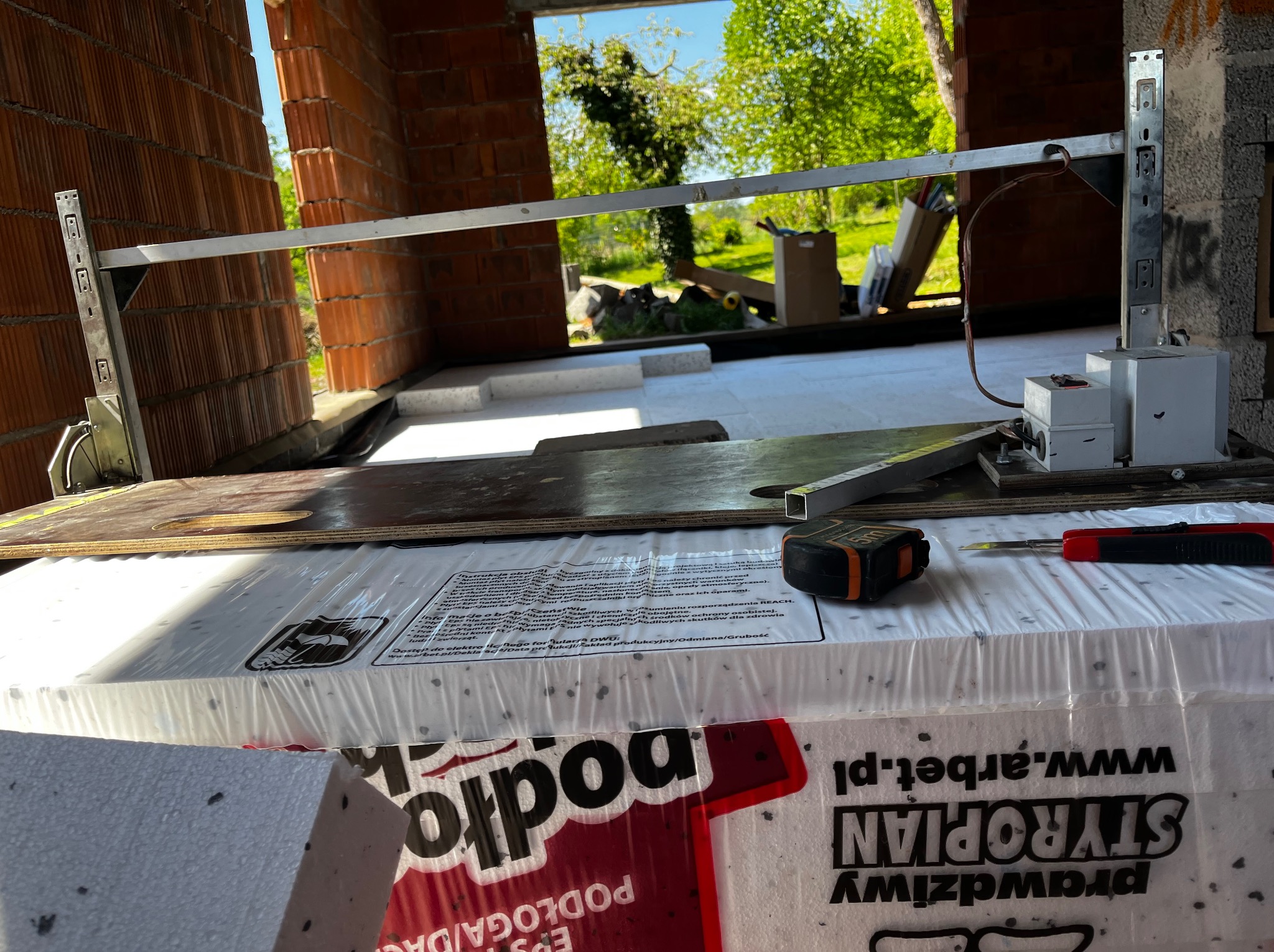 Przygotowanie do wylewki podłogi w nowo budowanym domu: styropian podłogowy, poziomica laserowa, miara zwijana i nożyk na tle ceglanej ściany i zieleni za oknem.