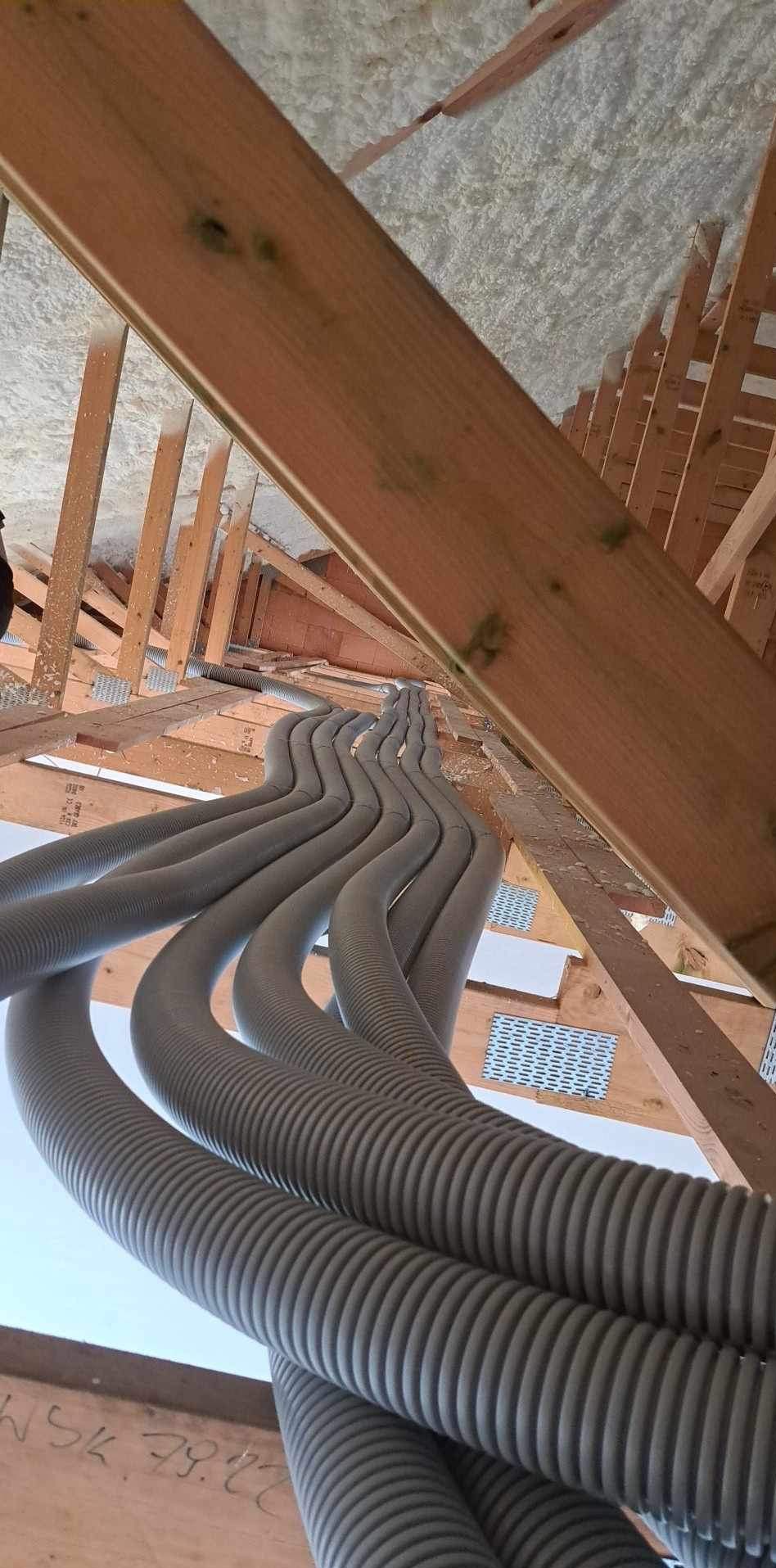 Rzędy szarych, karbowanych rur instalacyjnych ułożone równolegle w przestrzeni poddasza z drewnianą konstrukcją i izolacją piankową.
