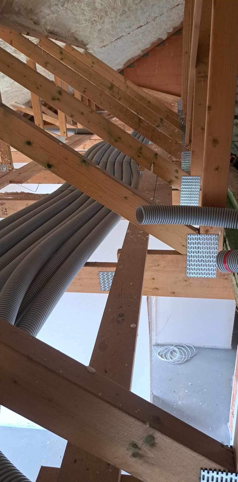 Szare, karbowane rury wentylacyjne ułożone wzdłuż drewnianej konstrukcji dachu, widoczne metalowe łączniki i fragment białej ściany w tle.