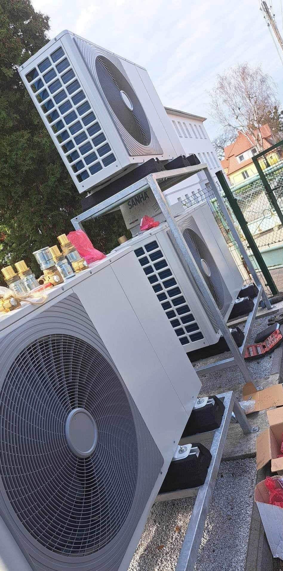 Trzy jednostki zewnętrzne klimatyzacji SANHA zamontowane na metalowej konstrukcji na zewnątrz budynku, widoczne elementy instalacyjne i kartony.