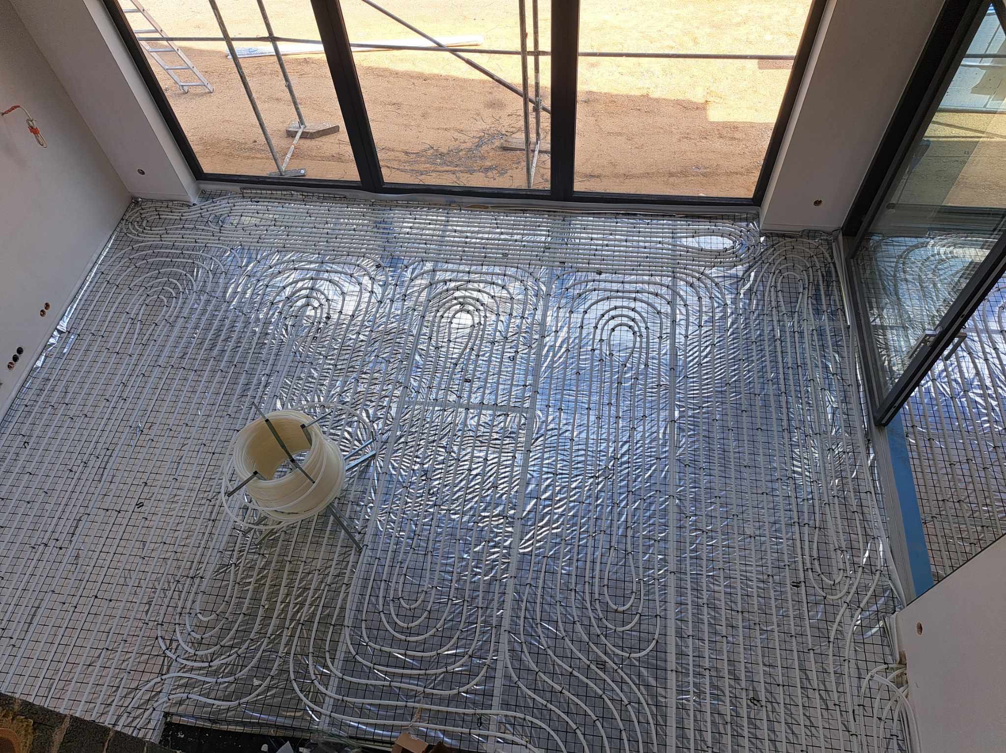 Widok z góry na instalację ogrzewania podłogowego z białych rurek ułożonych na srebrnej folii izolacyjnej i czarnej siatce, widoczne okno dachowe i zwój rur.