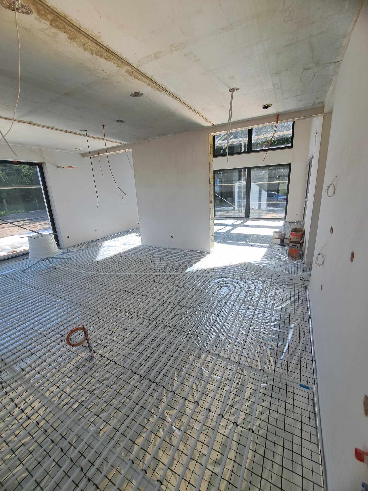 Układanie ogrzewania podłogowego w surowym stanie, rury grzewcze rozłożone na siatce, widoczne okna i surowy betonowy sufit z wystającymi przewodami elektrycznymi.