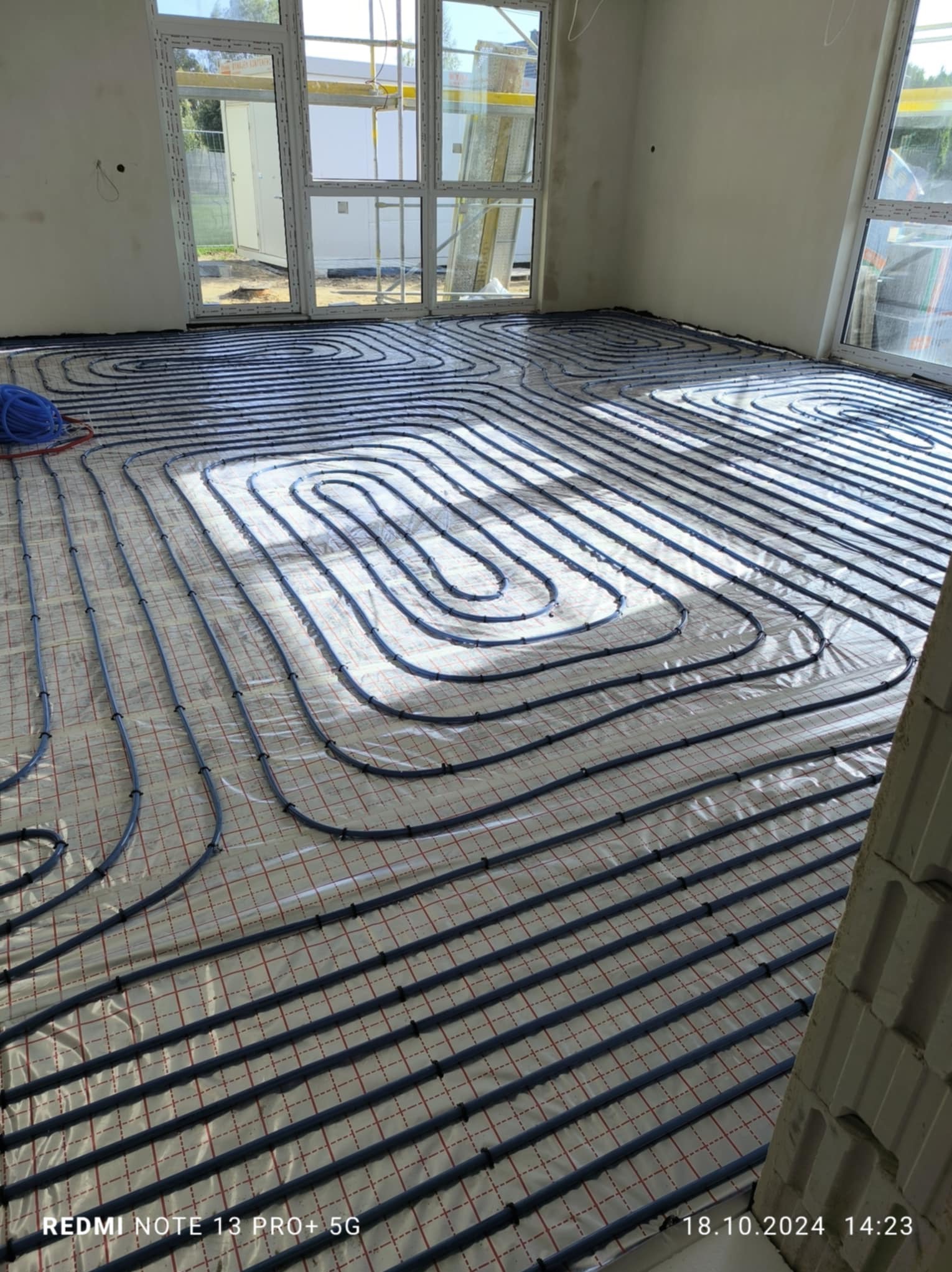 Wnętrze w trakcie prac instalacyjnych: rozłożone rury ogrzewania podłogowego na folii izolacyjnej z widocznymi oknami i fragmentem ściany z pustaków.