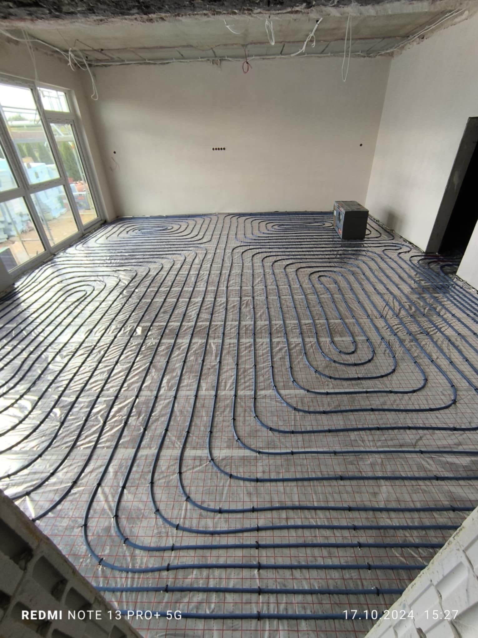 Niedokończona instalacja ogrzewania podłogowego z niebieskich rur na siatce, widok na surowy, jasny pokój z dużym oknem i betonowym sufitem.