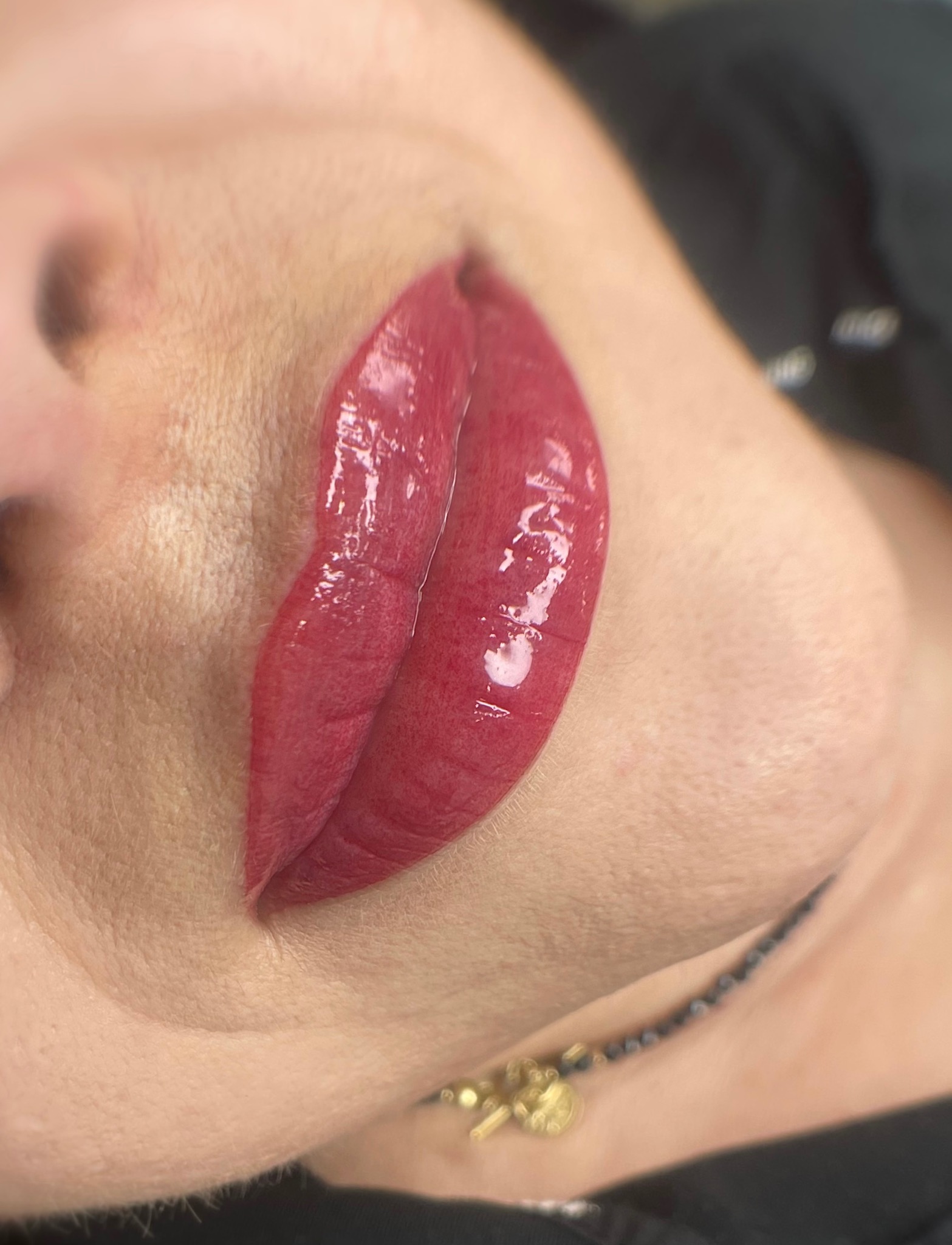 Zbliżenie na usta po zabiegu pigmentacji permanentnej, ukazujące intensywny czerwony kolor i błyszczące wykończenie, widoczna delikatna biżuteria na szyi modelki.