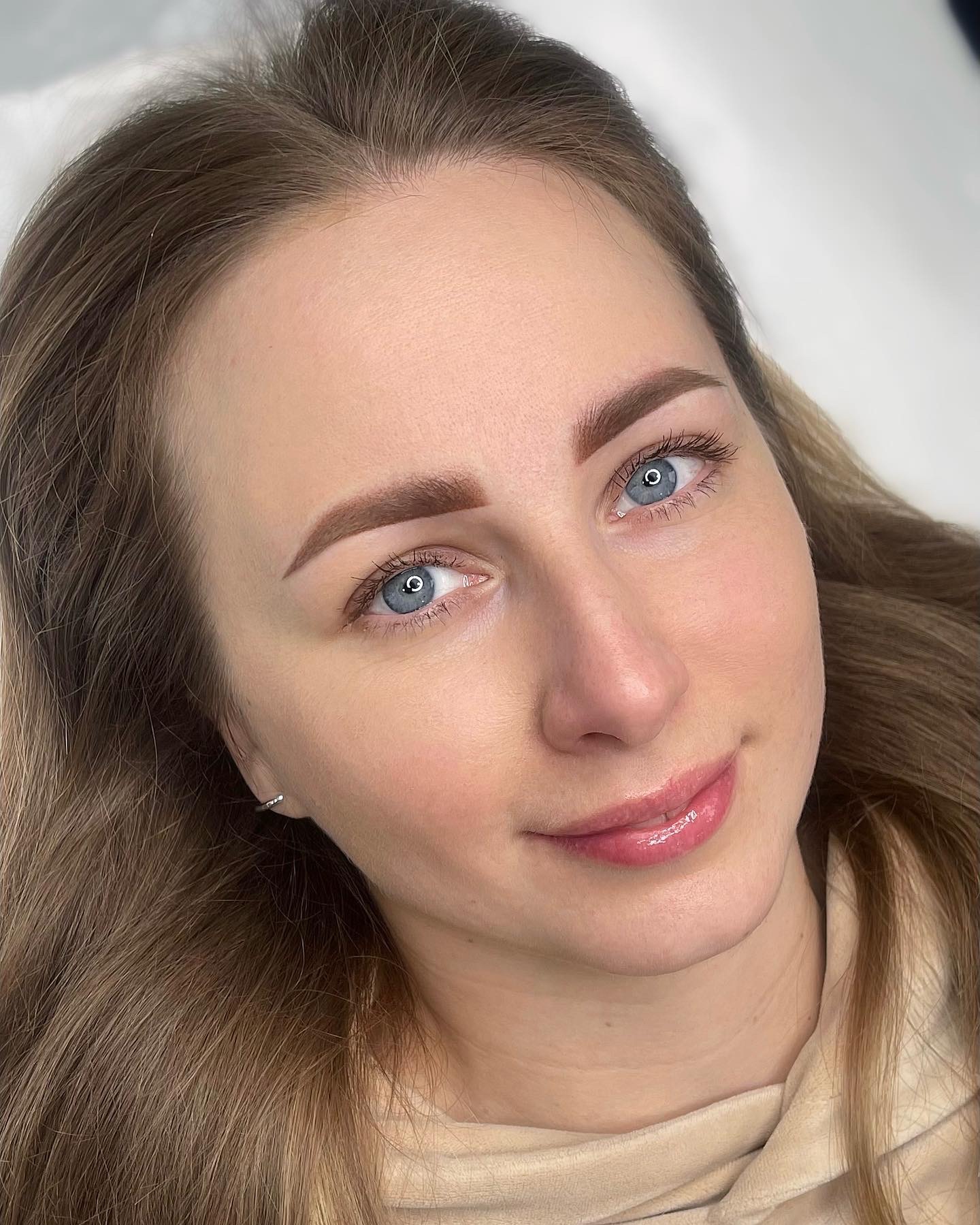 Portret kobiety z delikatnym makijażem permanentnym brwi w odcieniach brązu, podkreślającym naturalny kształt, z jasnymi, niebieskimi oczami i błyszczącymi ustami.
