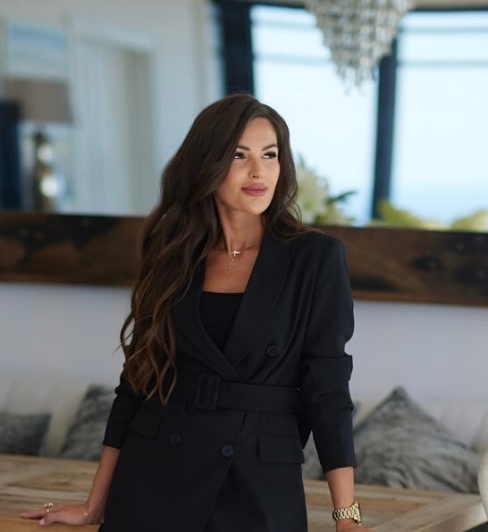 Portret kobiety z długimi brązowymi włosami, w czarnej marynarce z paskiem, subtelnym makijażu i złotym łańcuszku, pozującej w eleganckim wnętrzu z kryształowym żyrandolem.