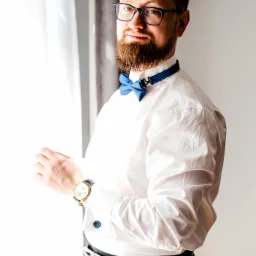 Mężczyzna z brodą i okularami, w białej koszuli i niebieskiej muszce, przygotowuje się do wyjścia, poprawiając mankiet koszuli z ozdobnymi spinkami i zegarek na skórzanym pasku, stojąc przy oknie.