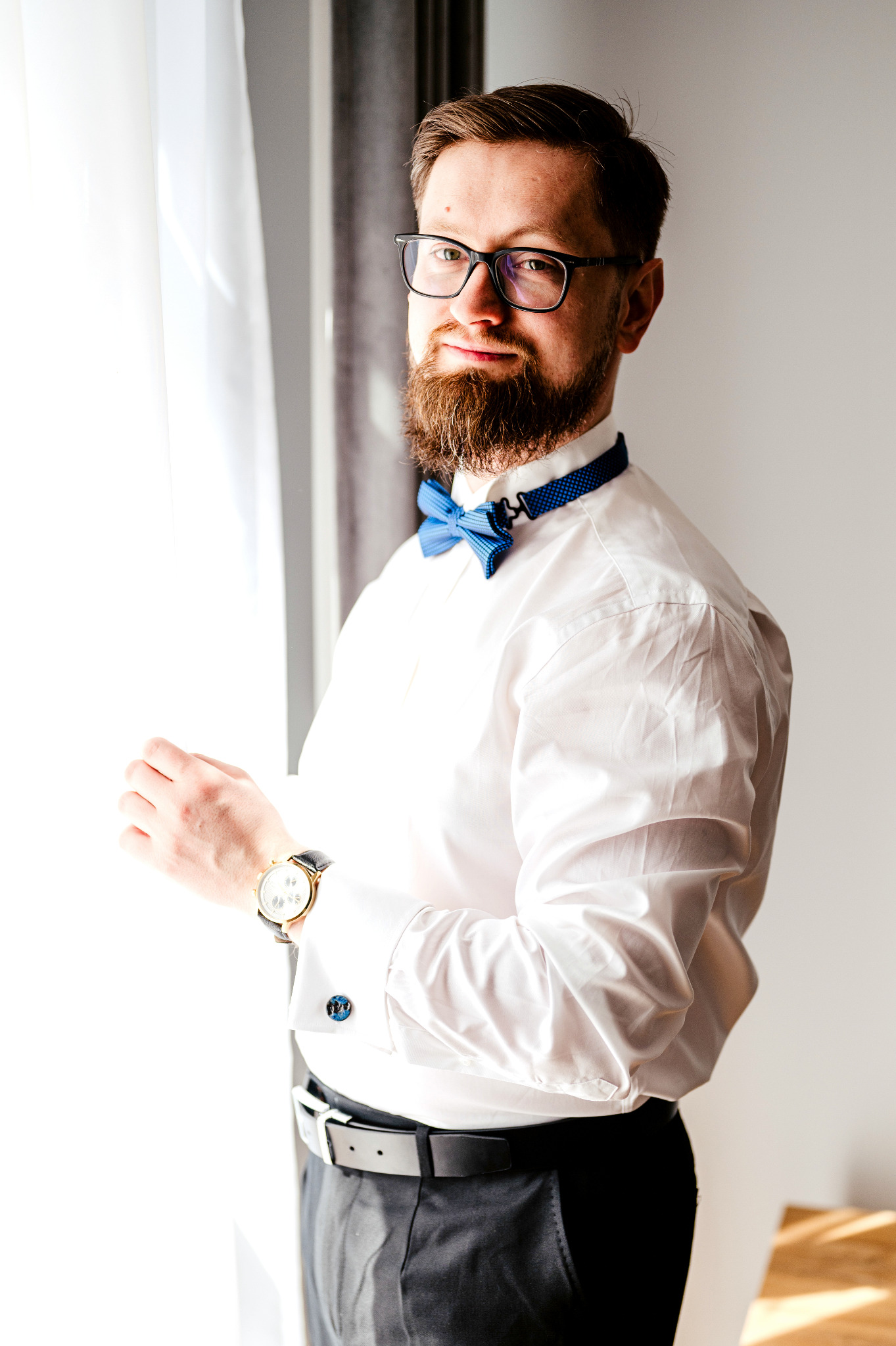 Mężczyzna z brodą i okularami, w białej koszuli i niebieskiej muszce, przygotowuje się do wyjścia, poprawiając mankiet koszuli z ozdobnymi spinkami i zegarek na skórzanym pasku, stojąc przy oknie.