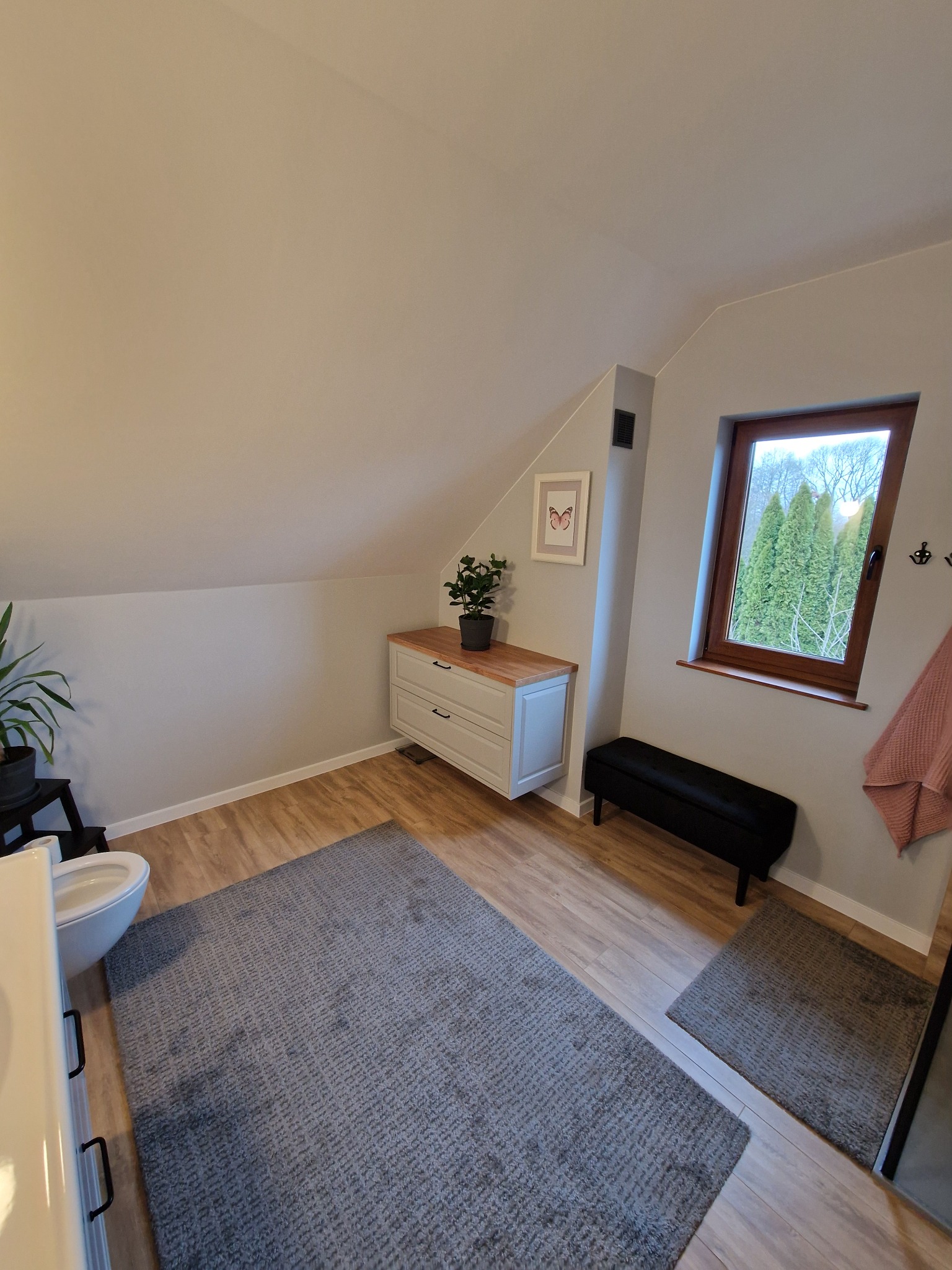 Jasna łazienka na poddaszu z drewnianą podłogą, szarym dywanem, komodą z rośliną, czarną ławką i oknem z widokiem na zieleń. Ukośne ściany i naturalne światło.