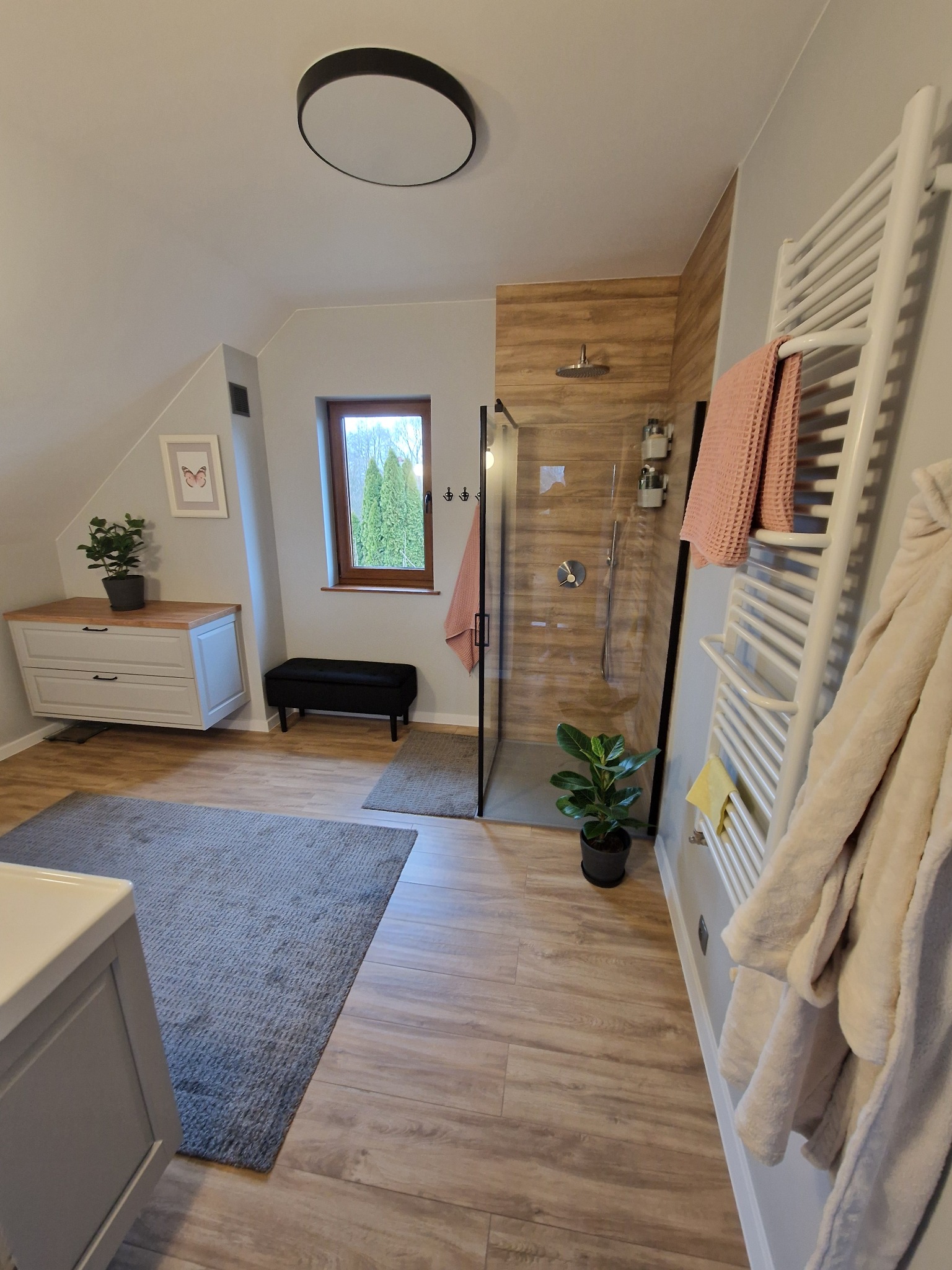 Jasna łazienka na poddaszu ze szklaną kabiną prysznicową z drewnopodobnymi panelami, grzejnikiem drabinkowym i szarym dywanem. Widok na okno i zieleń.