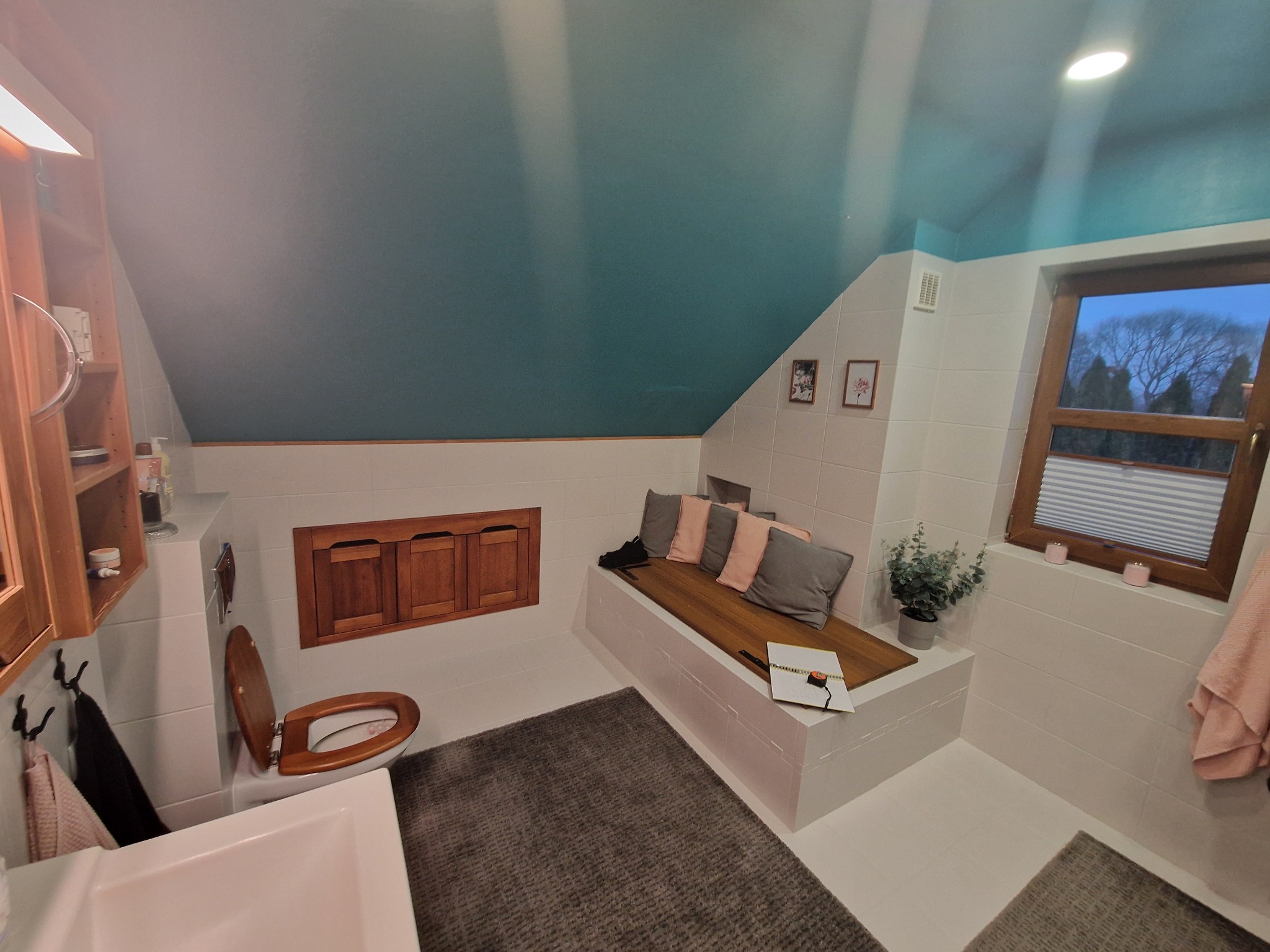 Wykończona łazienka na poddaszu z drewnianymi akcentami, turkusowym sufitem i oknem z widokiem na drzewa. Wnętrze doświetlone, z dywanem i siedziskiem z poduszkami.