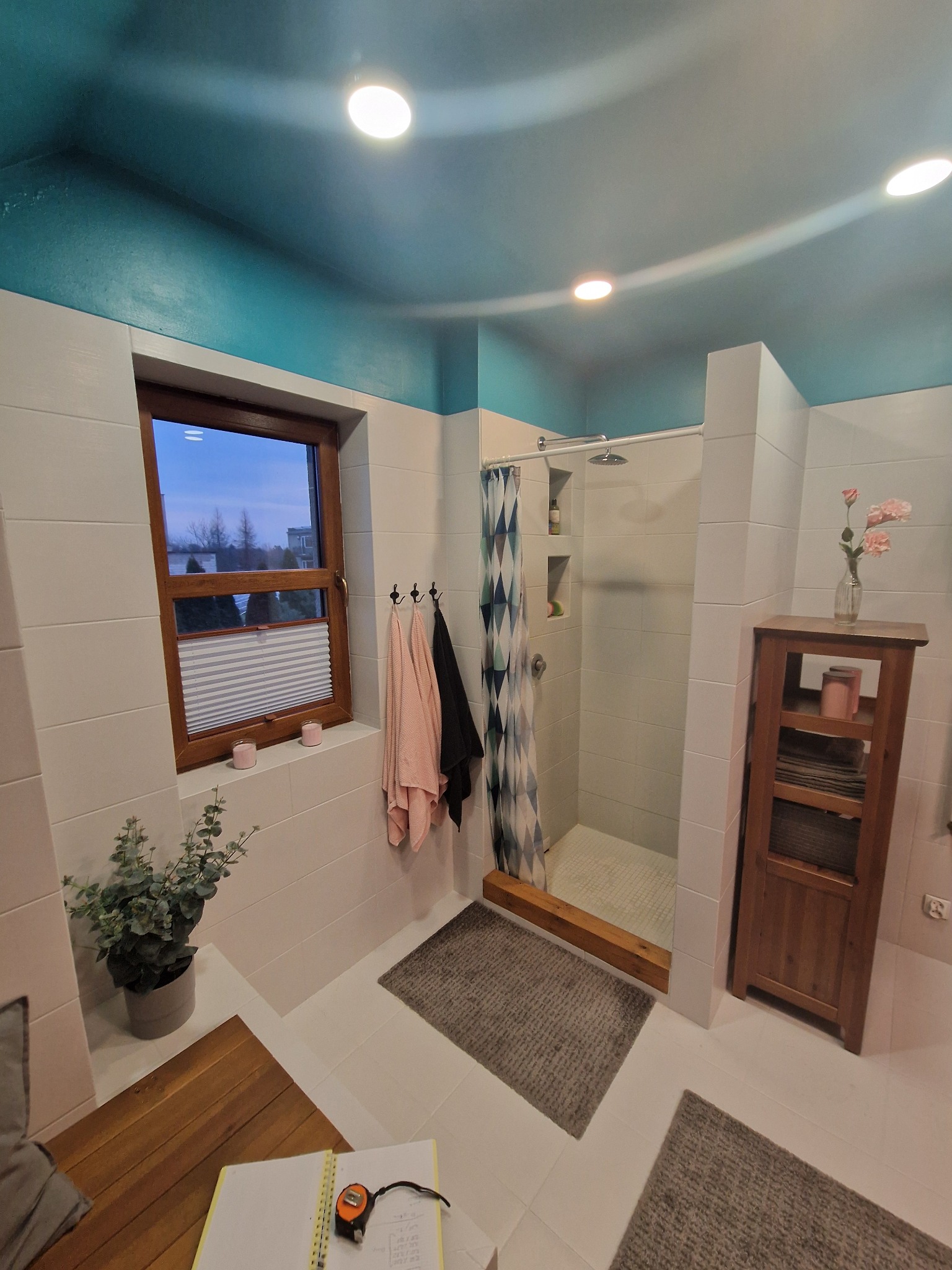 Wykończona łazienka w Ostaszewie z kabiną prysznicową, drewnianą szafką i oknem. Białe płytki, turkusowy sufit i dywaniki tworzą przytulne wnętrze.