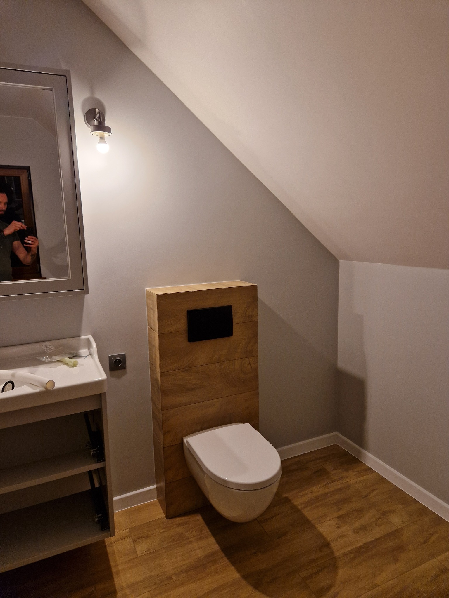 Nowoczesna łazienka na poddaszu: podwieszana toaleta z drewnianą obudową, umywalka z szafką i lustro. Ukośne ściany i drewnopodobna podłoga dodają wnętrzu charakteru.