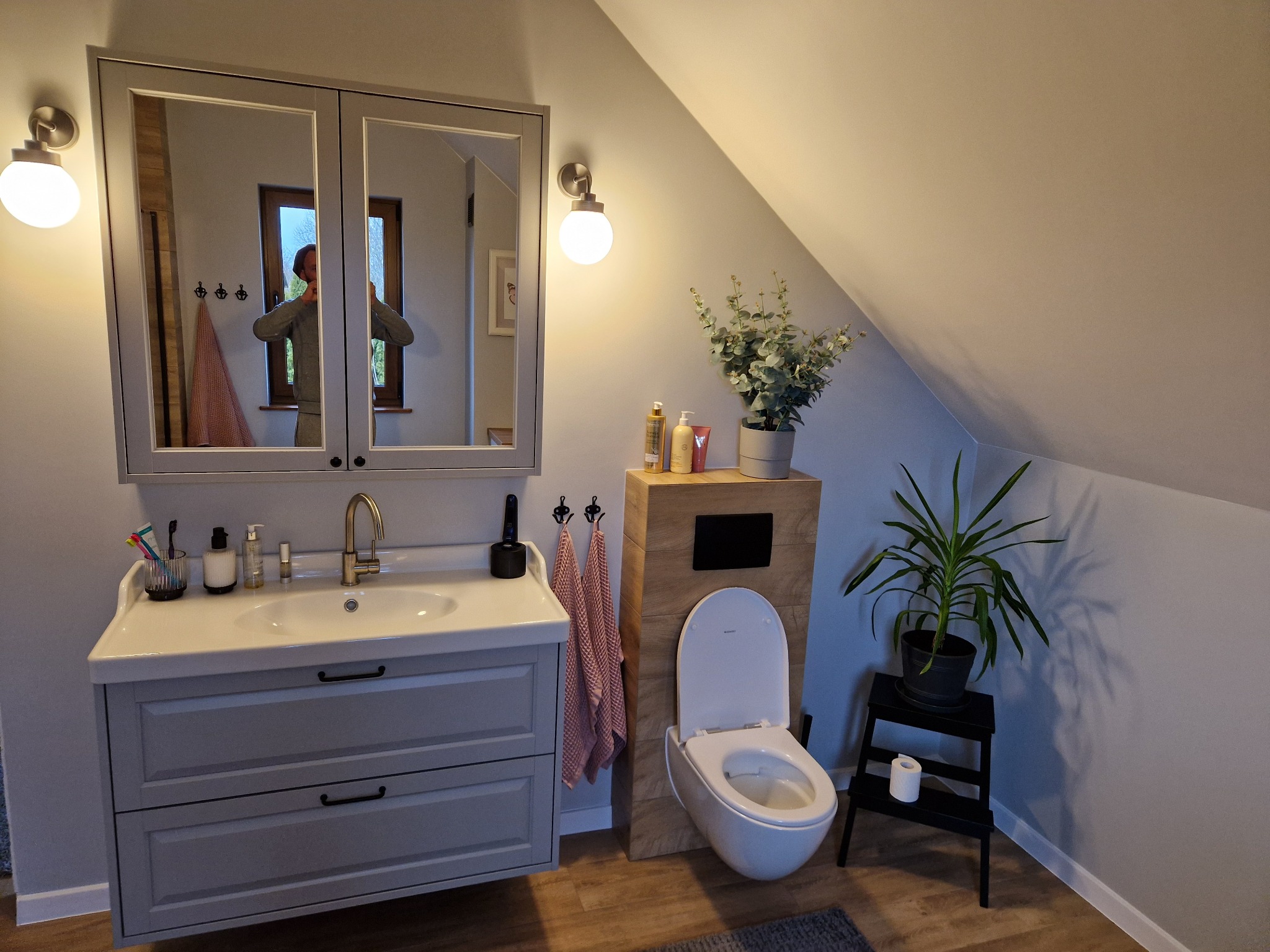 Jasna łazienka na poddaszu ze skośnym sufitem. Umywalka z szafką, toaleta w zabudowie, lustro z oświetleniem i rośliny doniczkowe tworzą przytulne wnętrze.