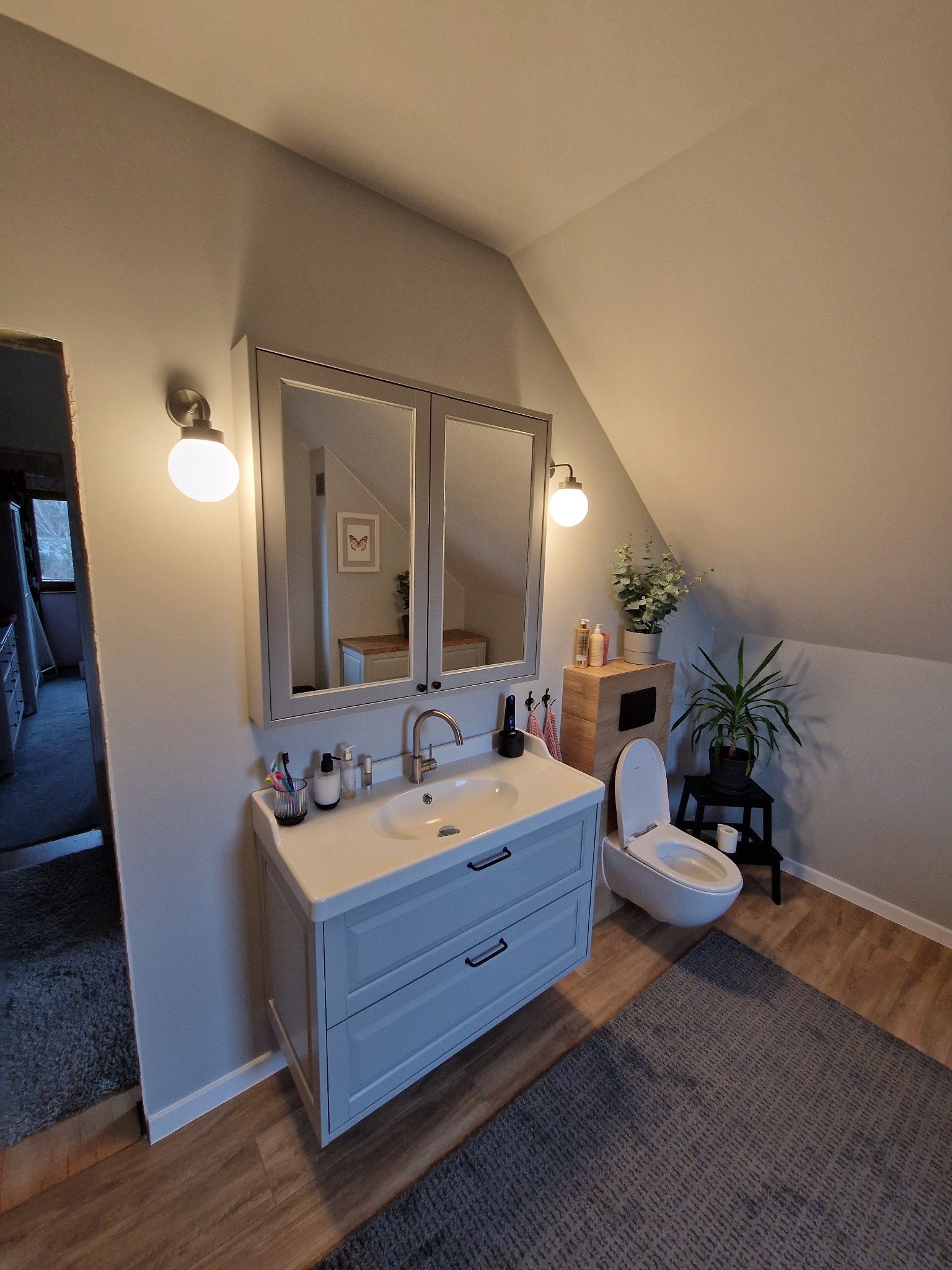 Jasna łazienka na poddaszu ze skośnym sufitem. Umywalka z szafką w stylu retro, lustro z oświetleniem, wisząca toaleta i rośliny doniczkowe tworzą przytulny klimat.