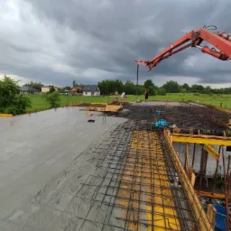 Wylewanie betonu na strop zbrojony, widoczna pompa do betonu, szalunki i pracujący robotnicy, w tle zabudowa jednorodzinna i pochmurne niebo.