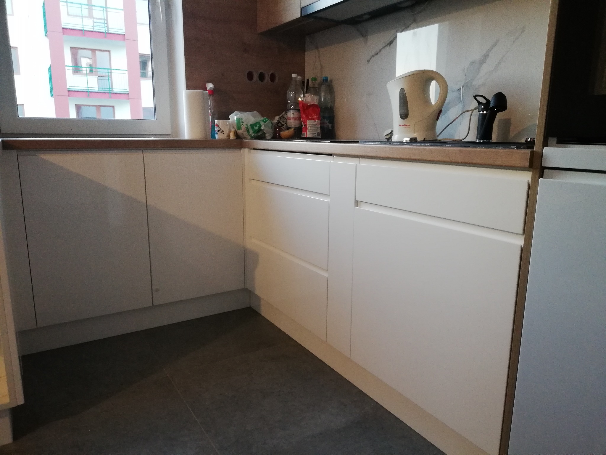 Biała, nowoczesna kuchnia na wymiar z drewnianym blatem, widok na okno z budynkiem mieszkalnym w tle, na blacie czajnik elektryczny i akcesoria kuchenne.