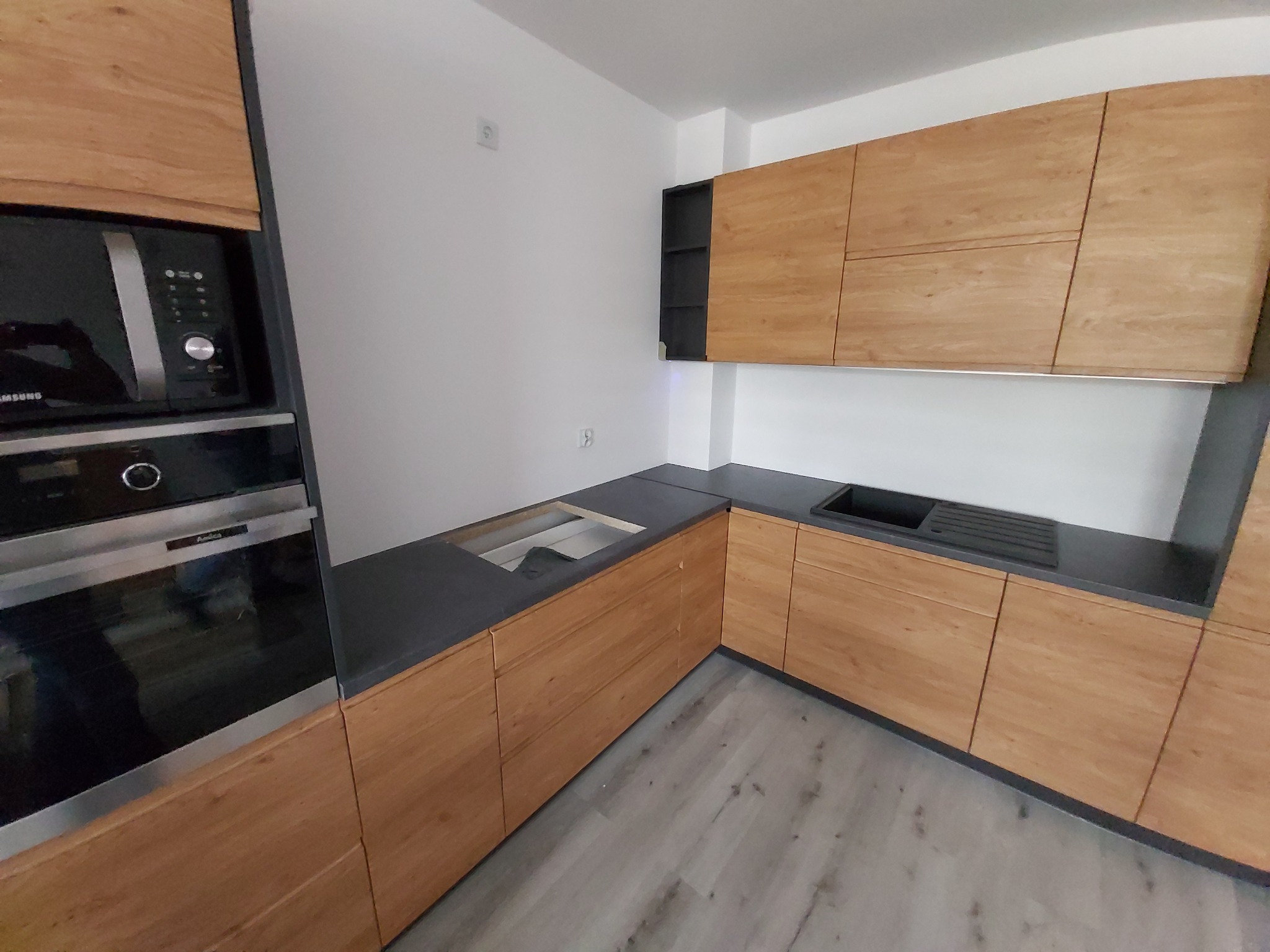 Kuchenna zabudowa z frontami w odcieniach drewna, szarym blatem i wbudowanym piekarnikiem oraz mikrofalówką, widok na narożnik i fragment podłogi.