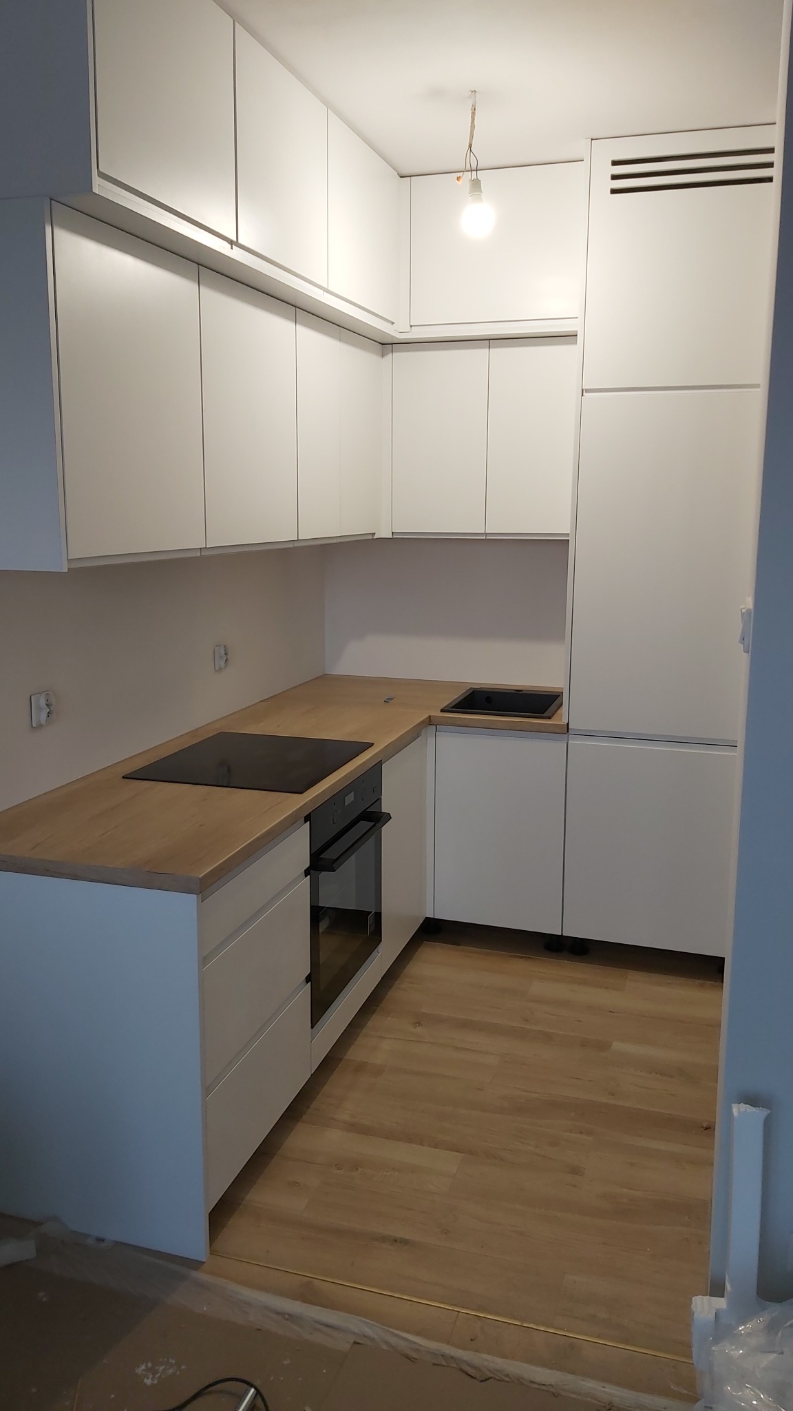 Nowoczesna, minimalistyczna kuchnia z białymi szafkami, drewnianym blatem, czarnym zlewem i płytą indukcyjną. Widoczna goła żarówka zamiast lampy i folia zabezpieczająca podłogę sugerują etap...