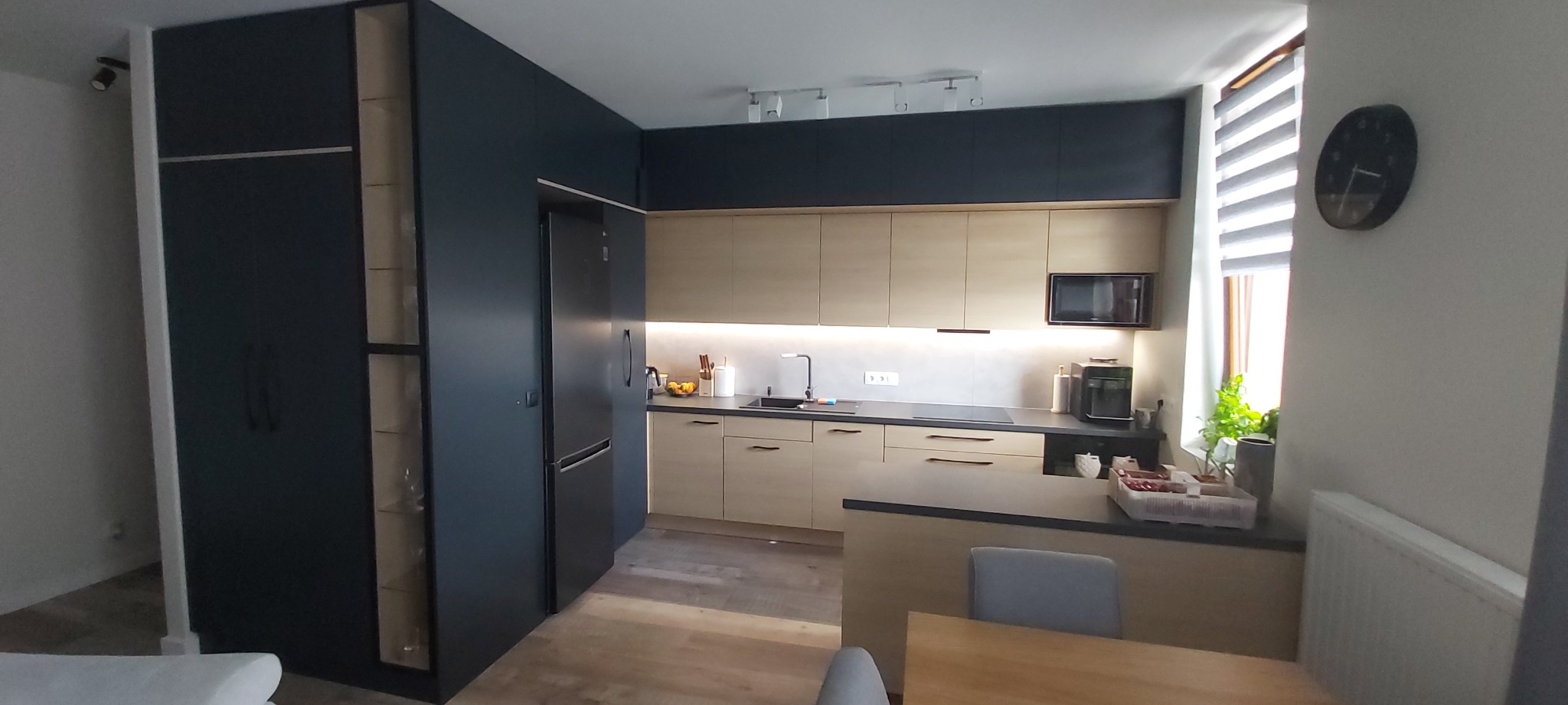 Nowoczesna kuchnia z zabudową w odcieniach beżu i granatu, z podświetleniem LED pod szafkami wiszącymi, widok na lodówkę i szafkę z przeszkleniami, zegar na ścianie w tle