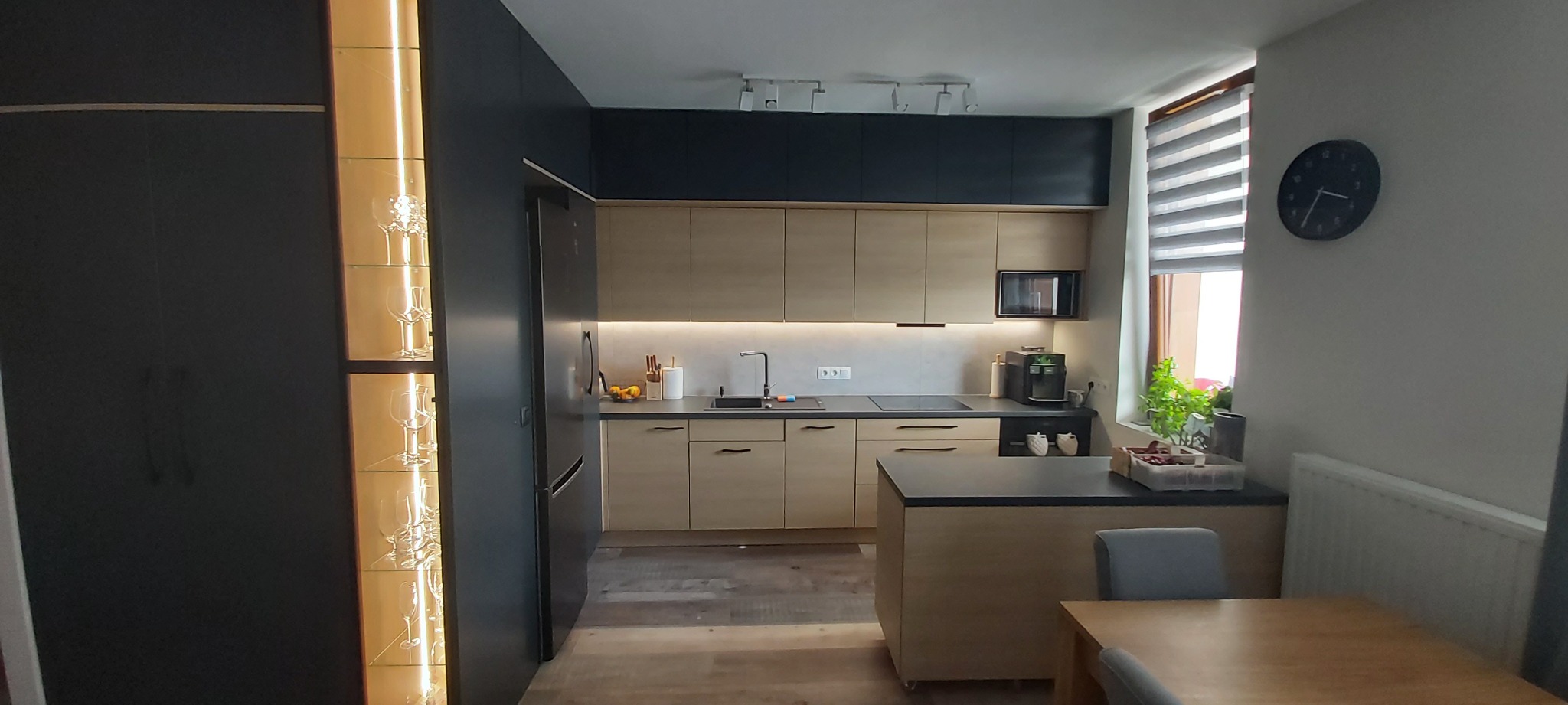 Nowoczesna kuchnia: beżowe szafki z czarnymi akcentami, oświetlona witryna z kieliszkami, czarny blat i sprzęt AGD, widok z jadalni.