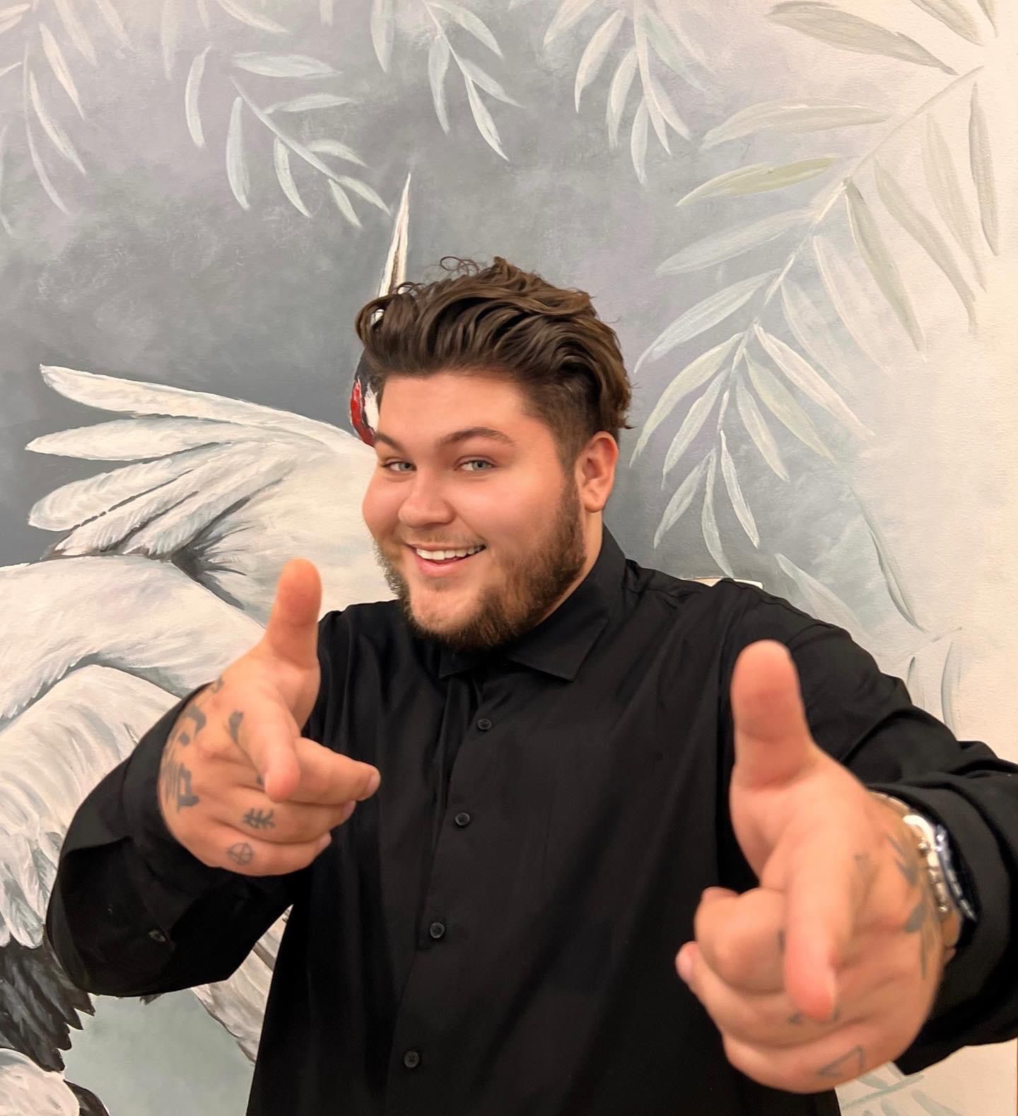 Uśmiechnięty mężczyzna w czarnej koszuli, z tatuażami na dłoniach, wskazujący palcami w stronę kamery, na tle ściany z malunkiem żurawia i liści.