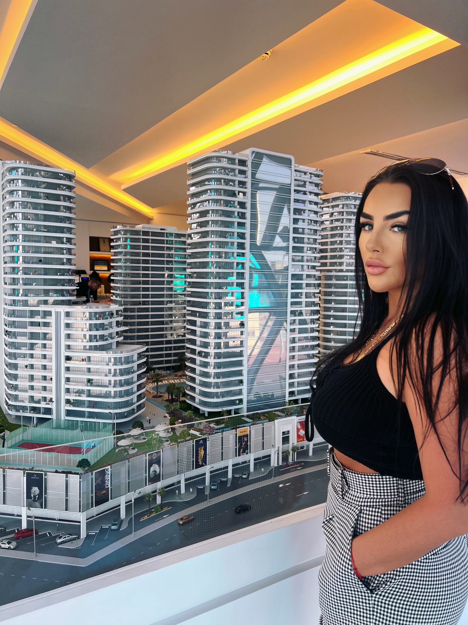 Kobieta pozuje obok makiety kompleksu apartamentów z kortem tenisowym, basenem i reklamami Rolex i Adidas.