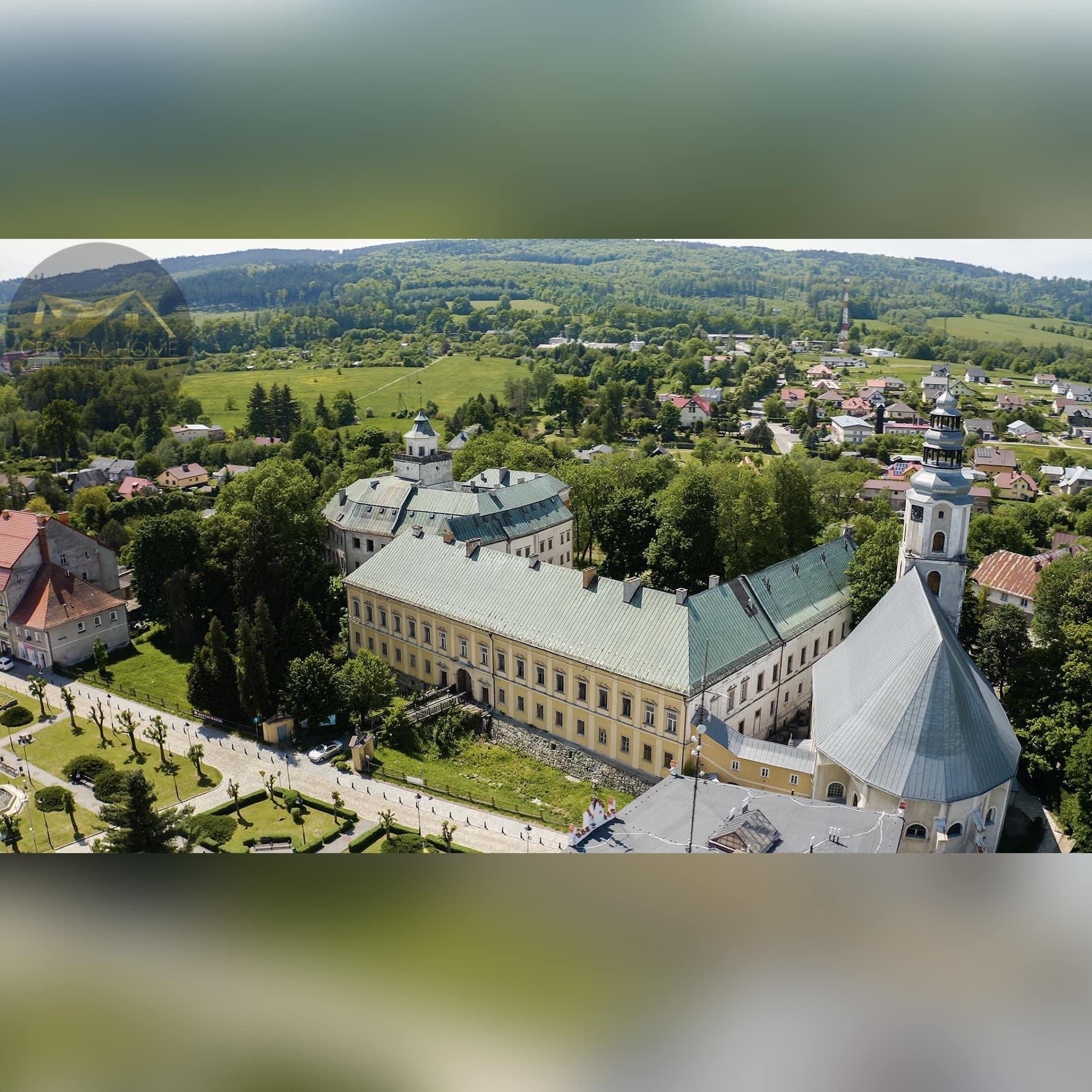 Panoramiczny widok z lotu ptaka na zabytkowy kompleks budynków z wieżą zegarową, otoczony zielenią i zabudową mieszkalną, w tle wzgórza i lasy, na pierwszym planie fragment dachu.