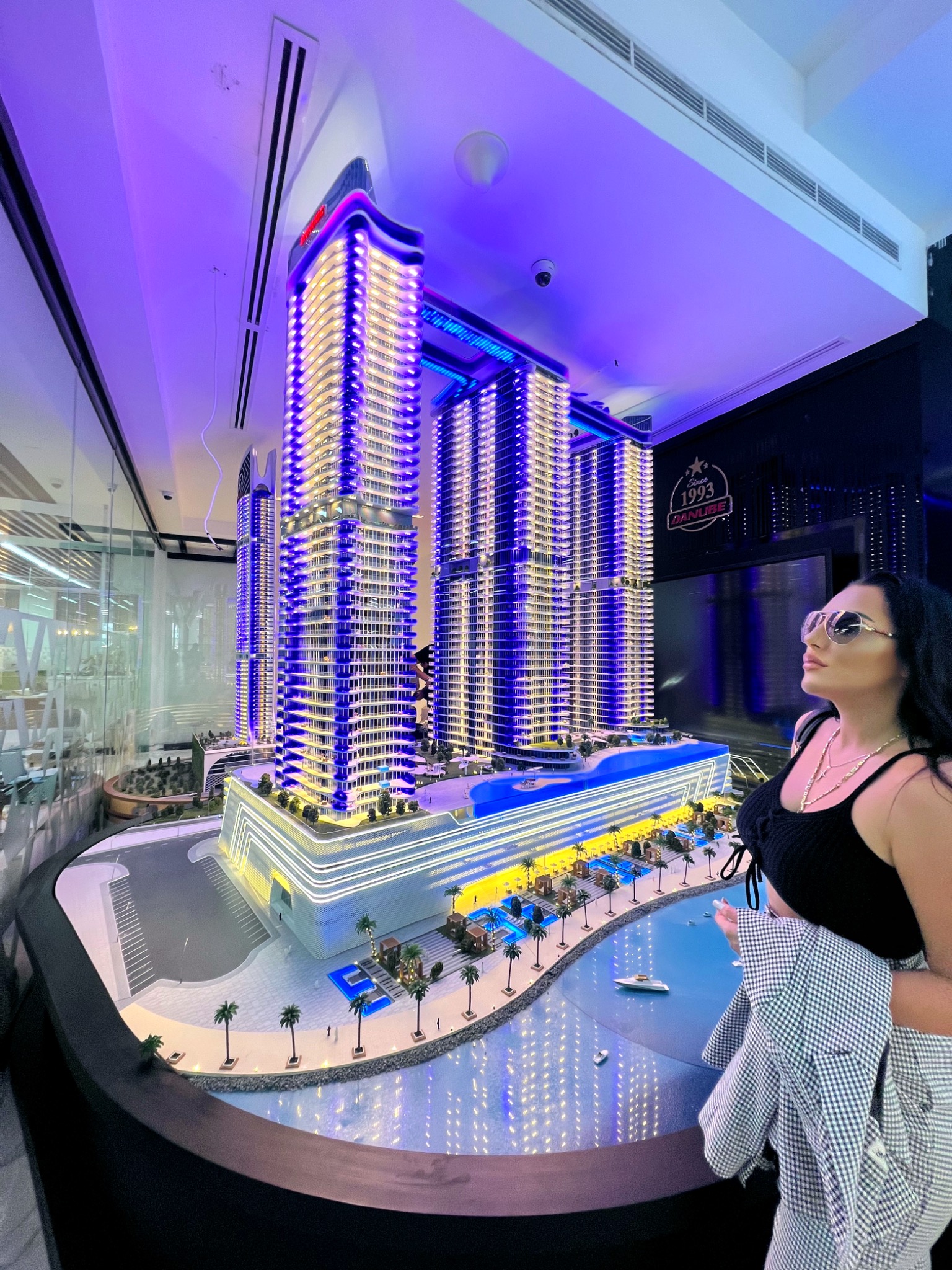 Makieta kompleksu budynków z oświetleniem, prezentowana przez kobietę w okularach przeciwsłonecznych, z widocznym logo '1993 Danube' w tle.