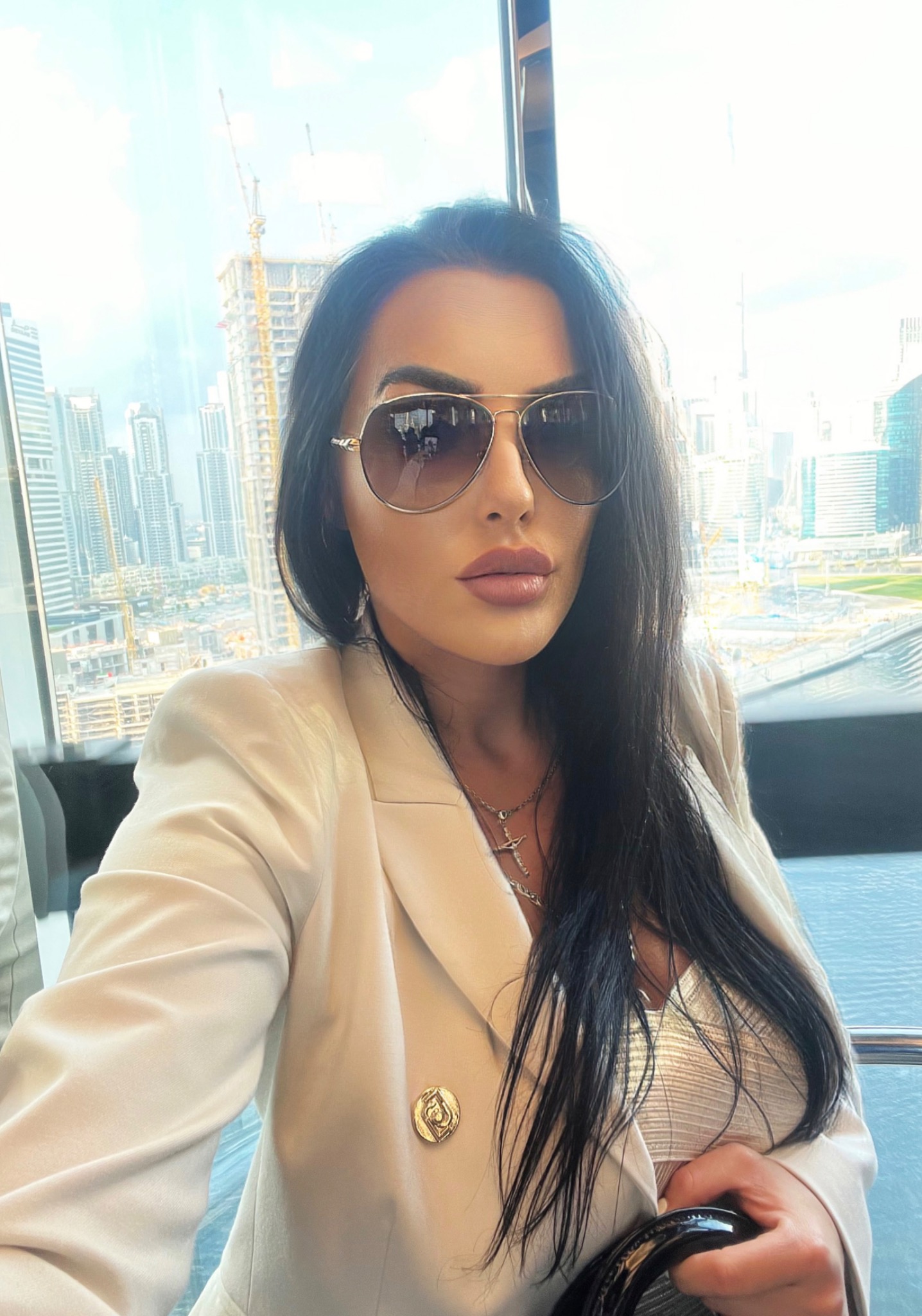 Kobieta w okularach przeciwsłonecznych i beżowym żakiecie pozuje na tle panoramy miasta z budową w Dubaju, widocznej przez okno.