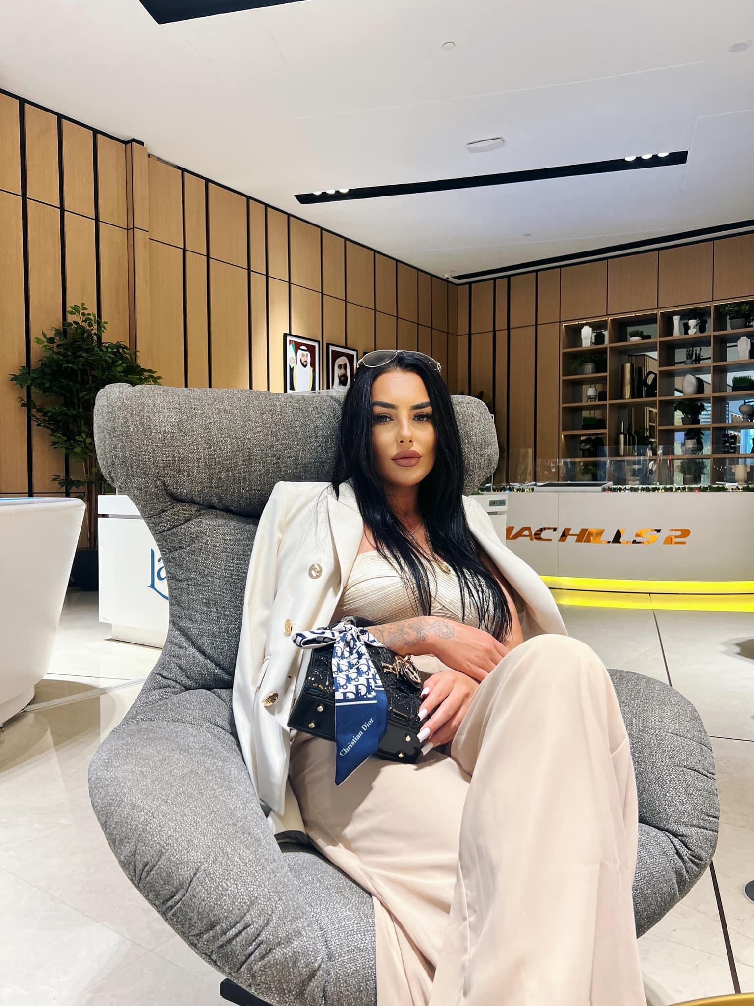 Kobieta w beżowym garniturze siedzi w szarym fotelu w nowoczesnym wnętrzu biura nieruchomości z ekspozycją modeli budynków w tle.