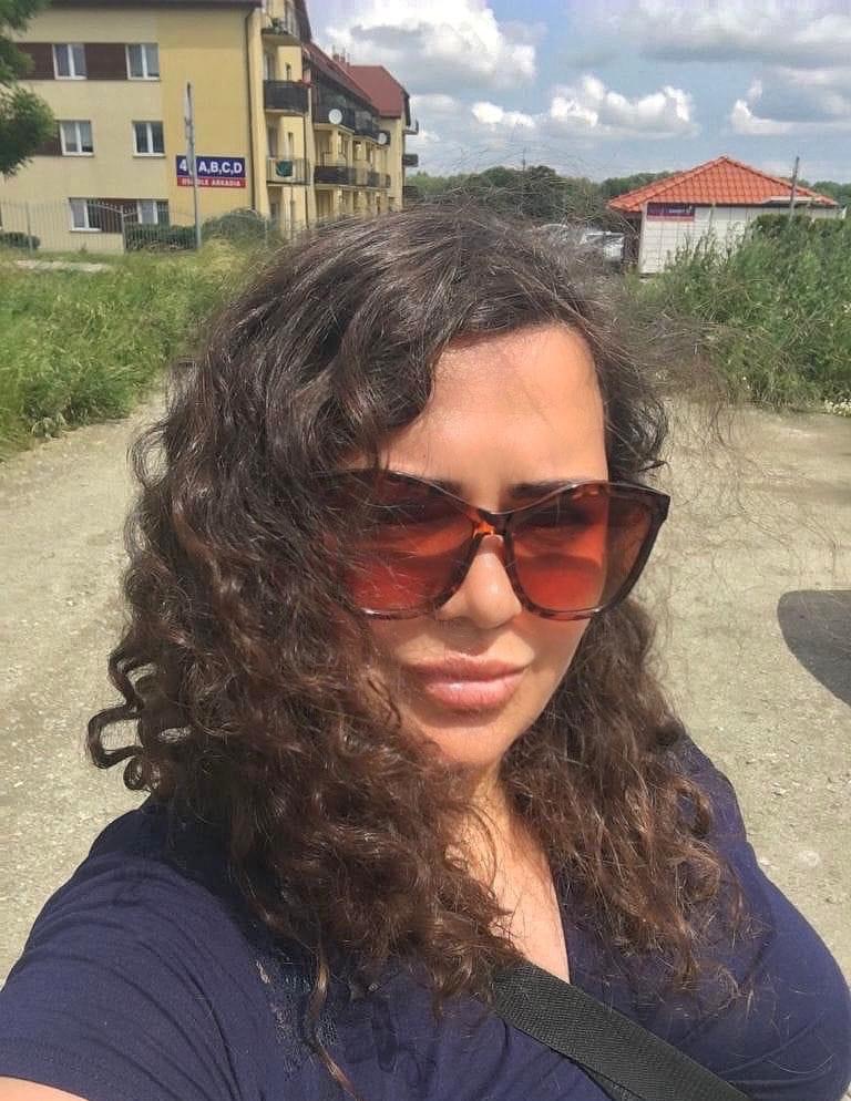 Portret kobiety w okularach przeciwsłonecznych na tle budynku mieszkalnego z tabliczką adresową i zieleni