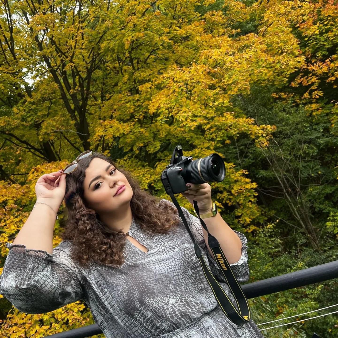 Kobieta z kręconymi włosami trzyma aparat Nikon na tle jesiennych drzew, z ręką unoszącą okulary przeciwsłoneczne, ubrana w bluzkę imitującą skórę krokodyla.