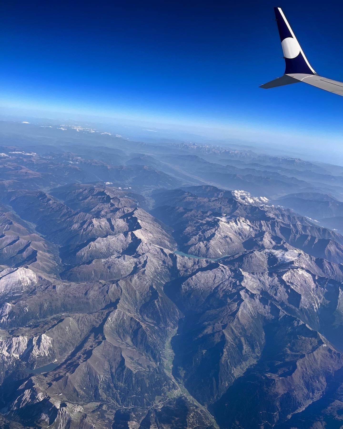 Widok z okna samolotu na Alpy z błękitnym jeziorem i fragmentem skrzydła samolotu w prawym górnym rogu.