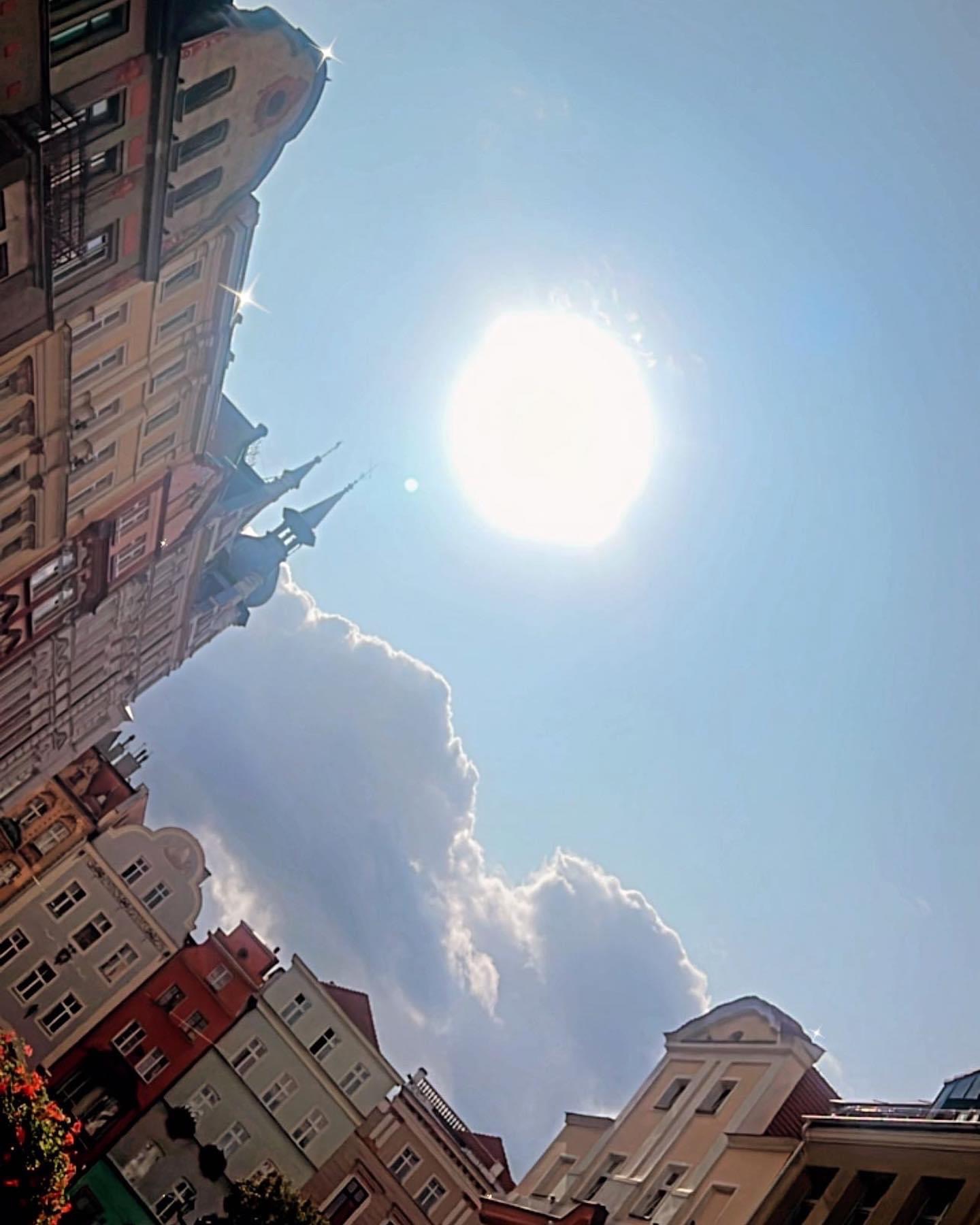 Kolorowe kamienice w historycznej zabudowie miejskiej, widziane z dołu pod ostrym kątem, na tle błękitnego nieba z jasnym słońcem i białymi chmurami.